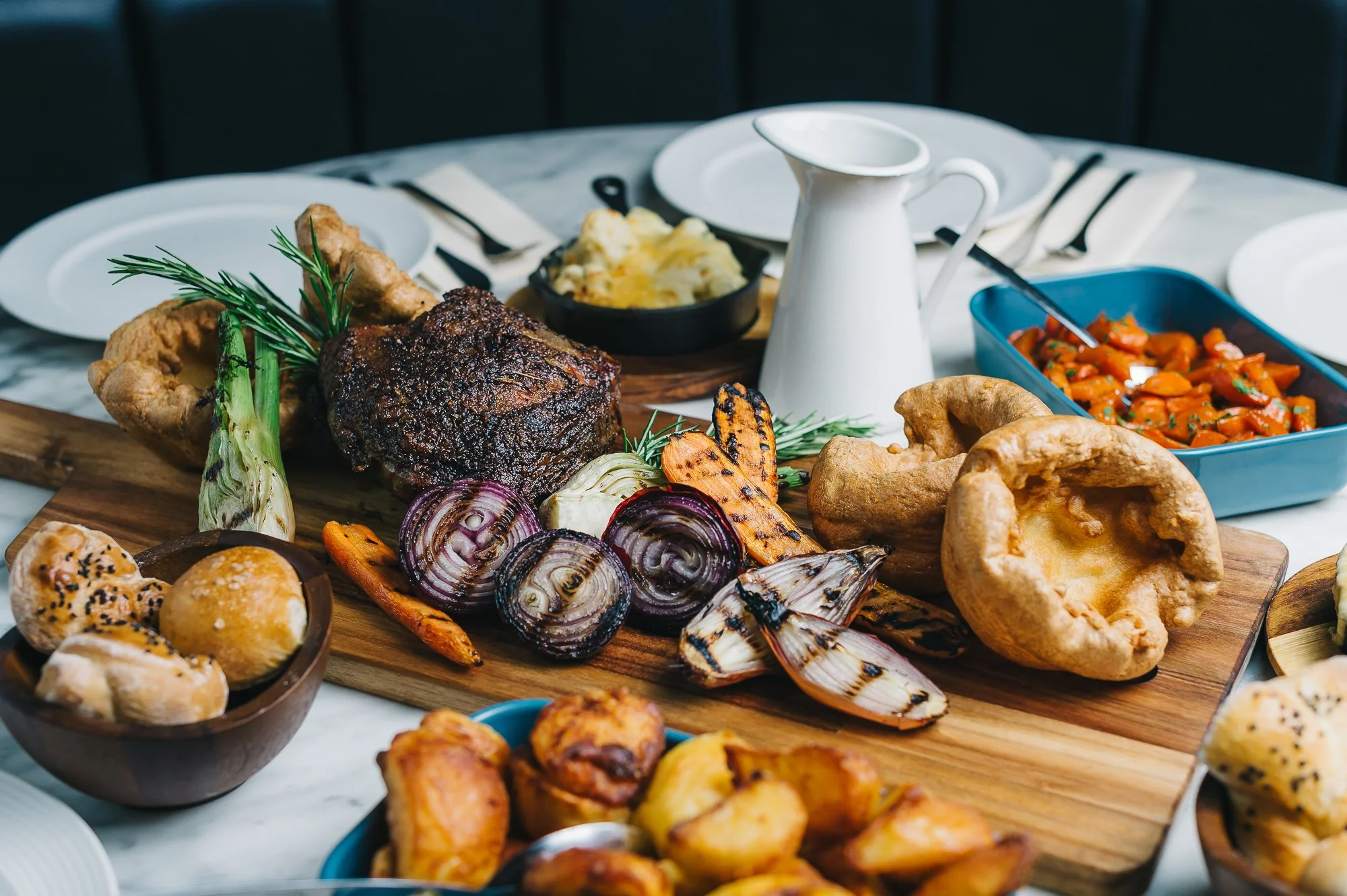 A variety of grilled and roasted vegetables and meats served on a wooden platter, including onion, carrots, and a large piece of cooked beef, with sides of mashed potatoes, baked potatoes, and a spicy vegetable dish, set on a white table with plates 