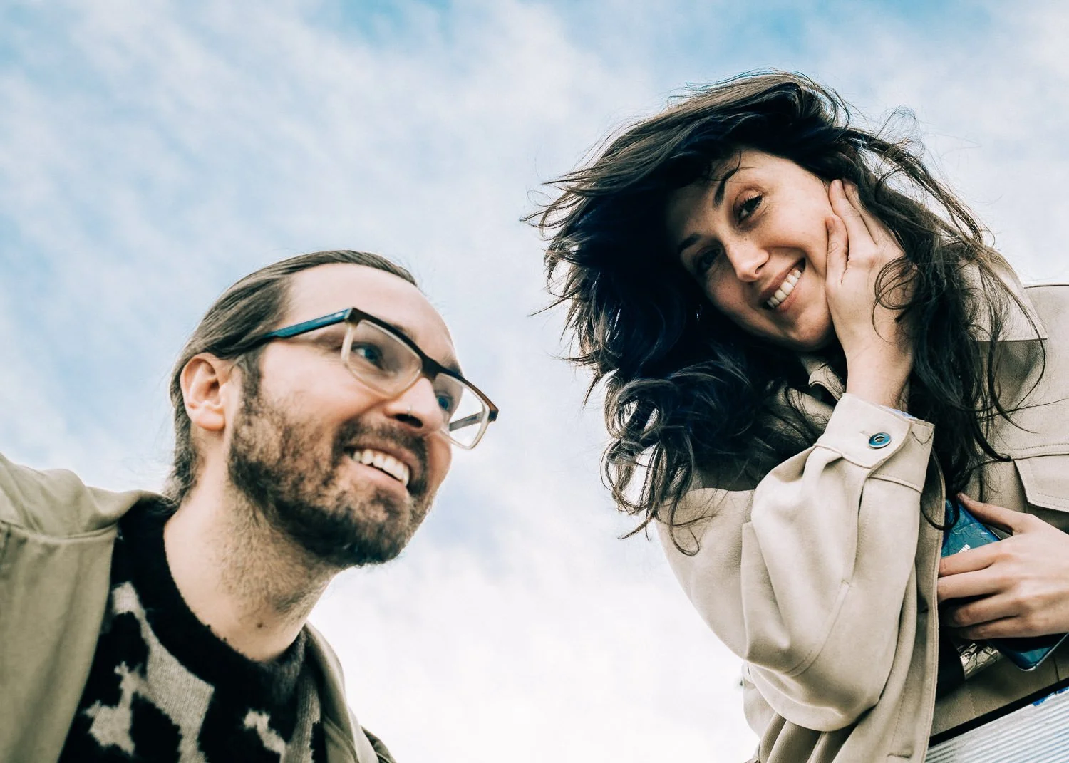 A man with glasses and a woman with dark hair smiling outdoors against a cloudy sky.