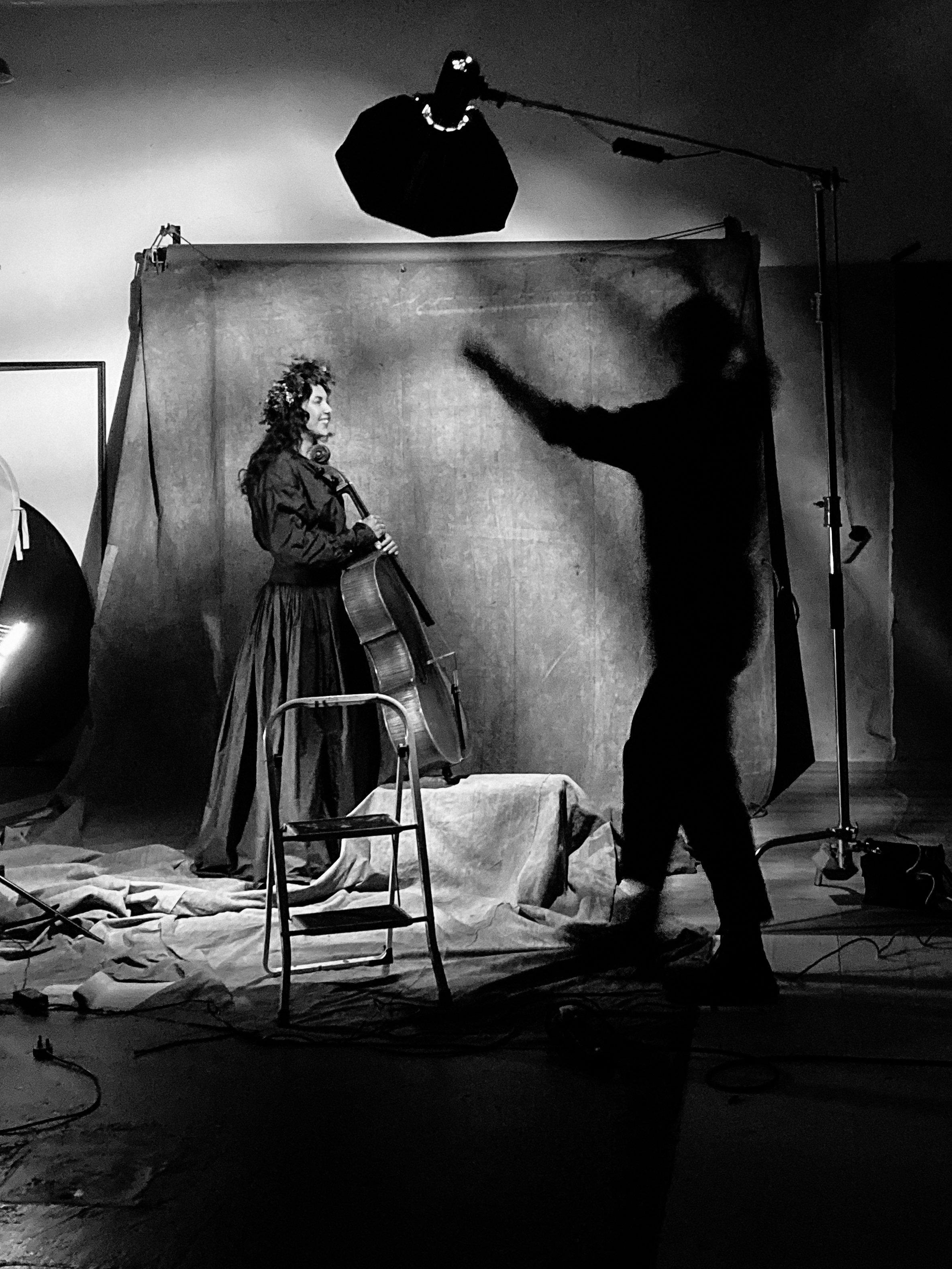 Black and white photo of a woman holding a guitar, smiling, while a man creates a shadow silhouette of her with his hand against a backdrop, in a photography studio.