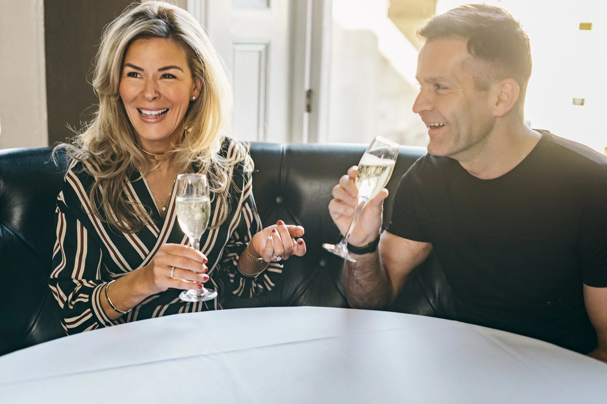 A woman and man sitting on a black leather couch, smiling and enjoying drinks in a well-lit room.
