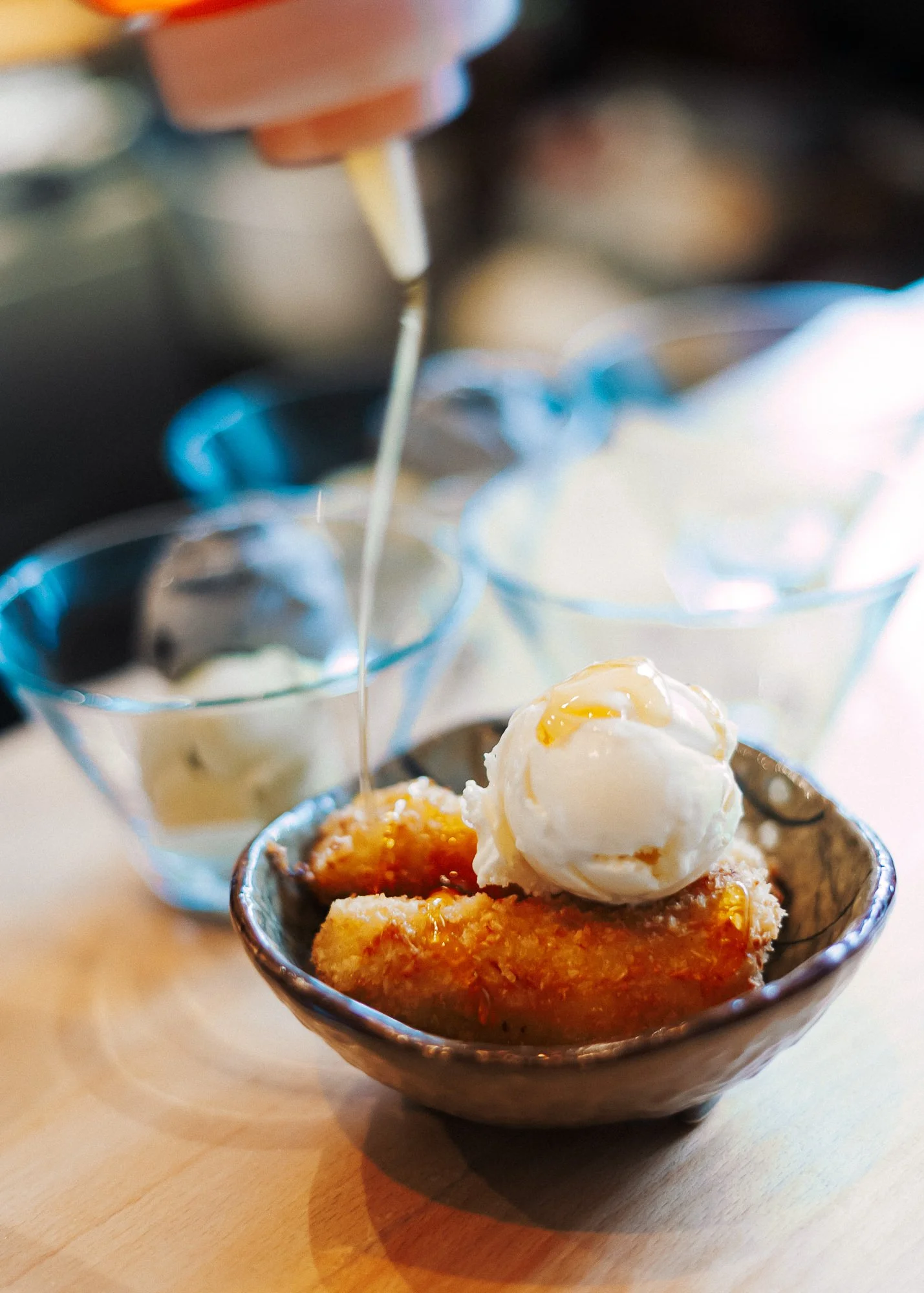 A dessert bowl with a slice of fruit pie topped with a scoop of vanilla ice cream, with syrup being poured over it. In the background, small bowls of ice cream are visible.