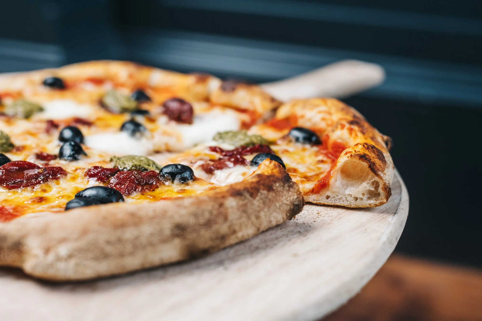 Close-up of a pizza with black olives, green pesto, and melted cheese on a wooden serving board.