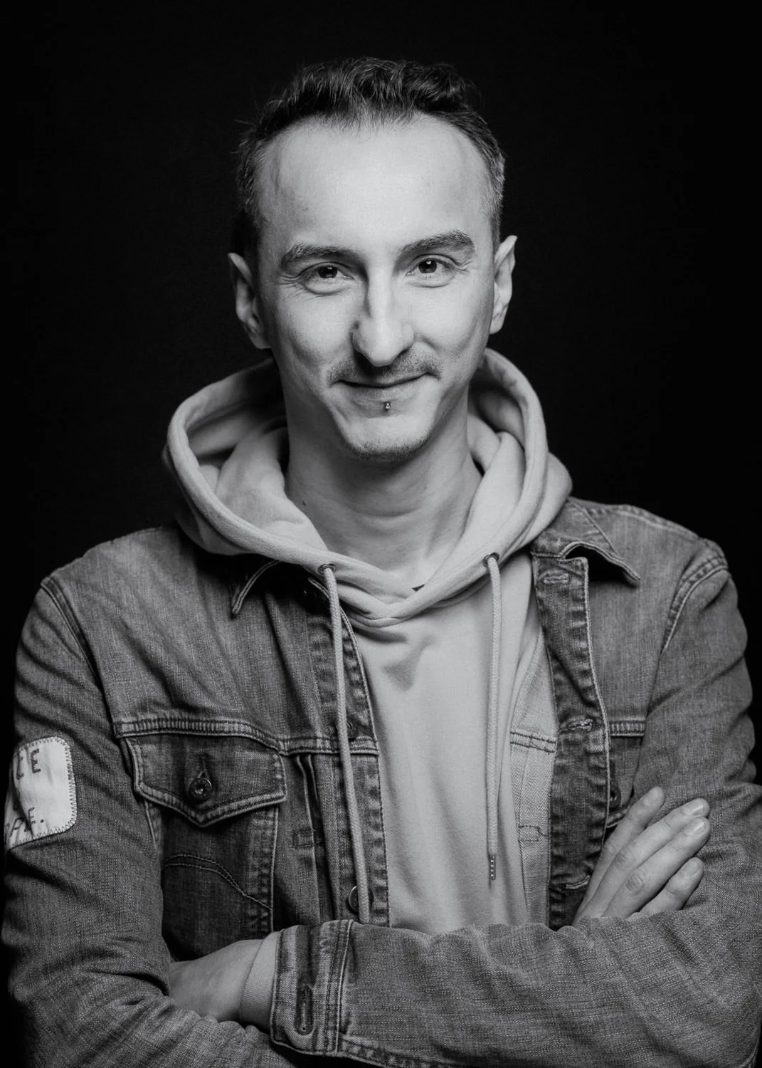 Black and white portrait of a young man with short hair, wearing a hoodie and a denim jacket, smiling with arms crossed.
