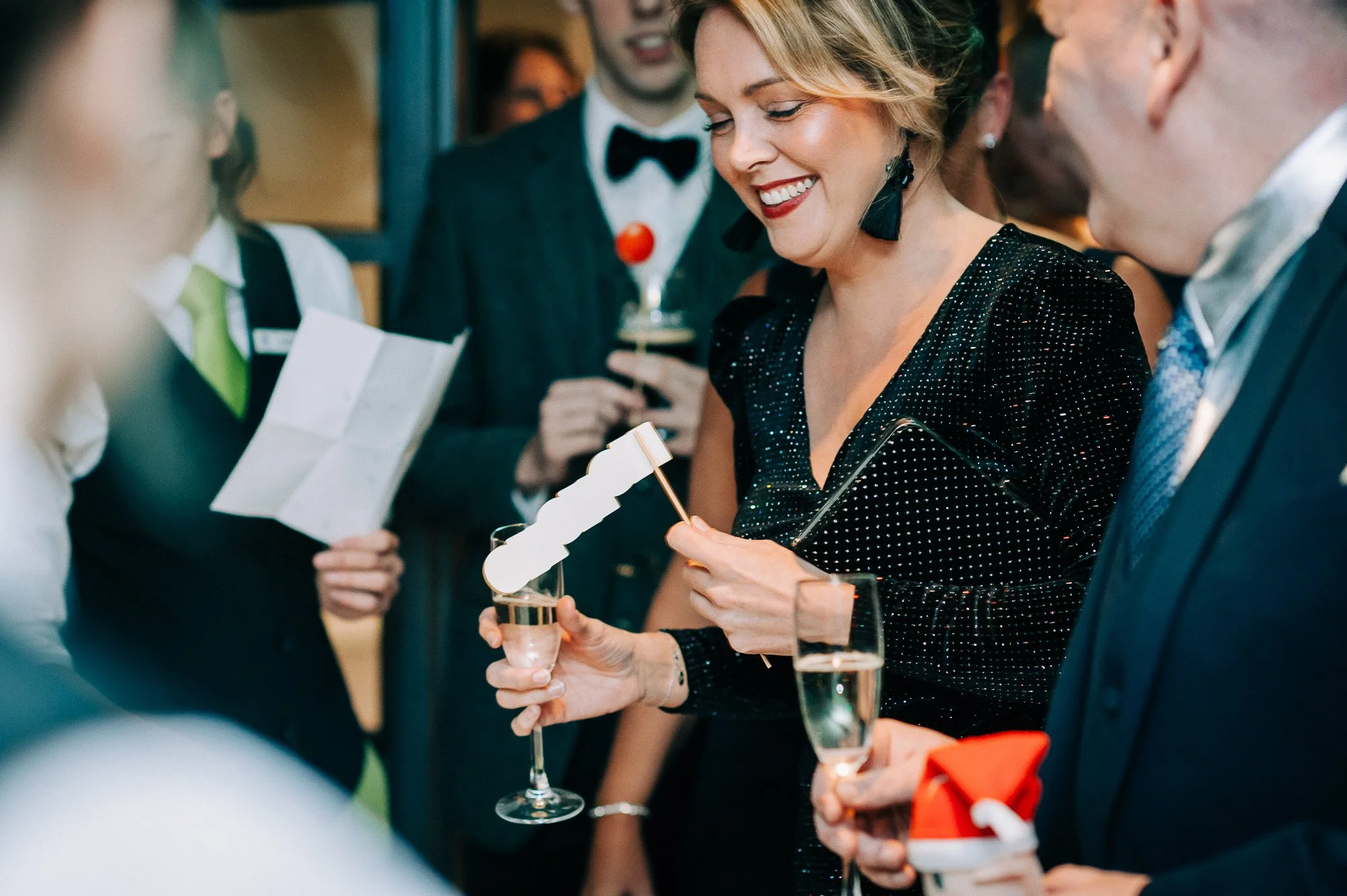 A woman in a black, sparkly dress smiling at a celebration party, holding a glass of champagne and a small flag, surrounded by well-dressed guests.