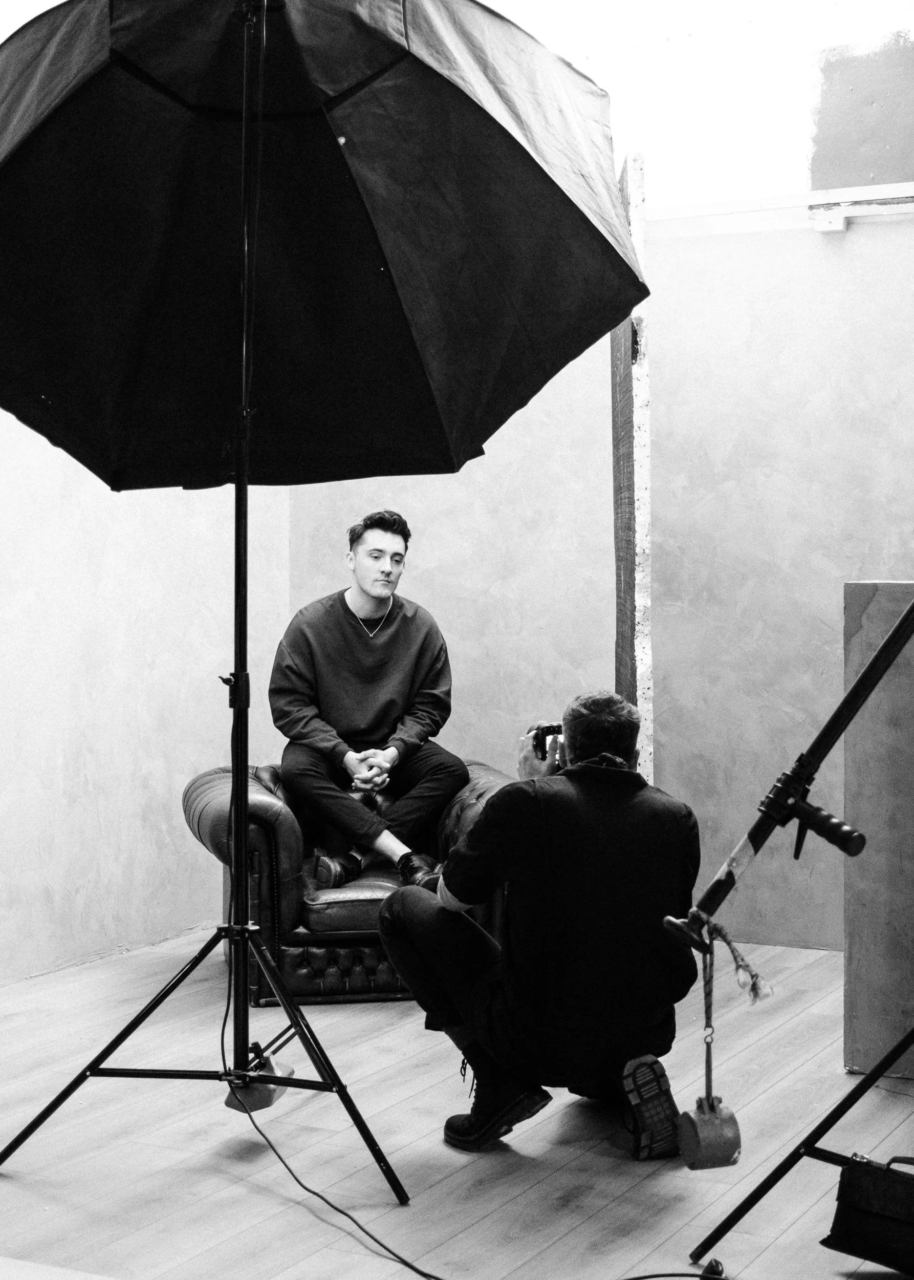 A young man sits on an armchair during a photo or video shoot in a studio with plain walls, with photographer kneeling in front of him taking pictures and large studio lights and umbrella for lighting.