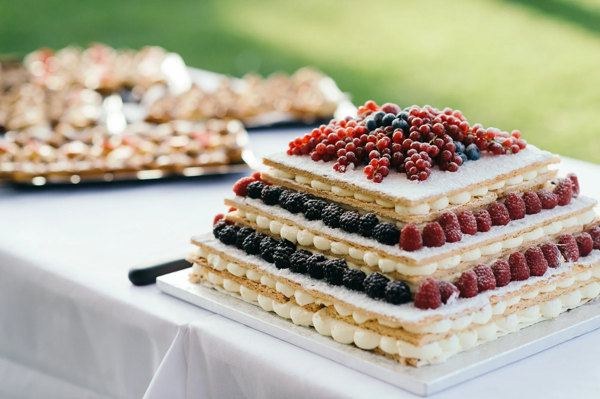 A layered rectangular mille feuille cake decorated with red and black berries, with each layer separated by white cream, sitting on a white tablecloth outdoors.
