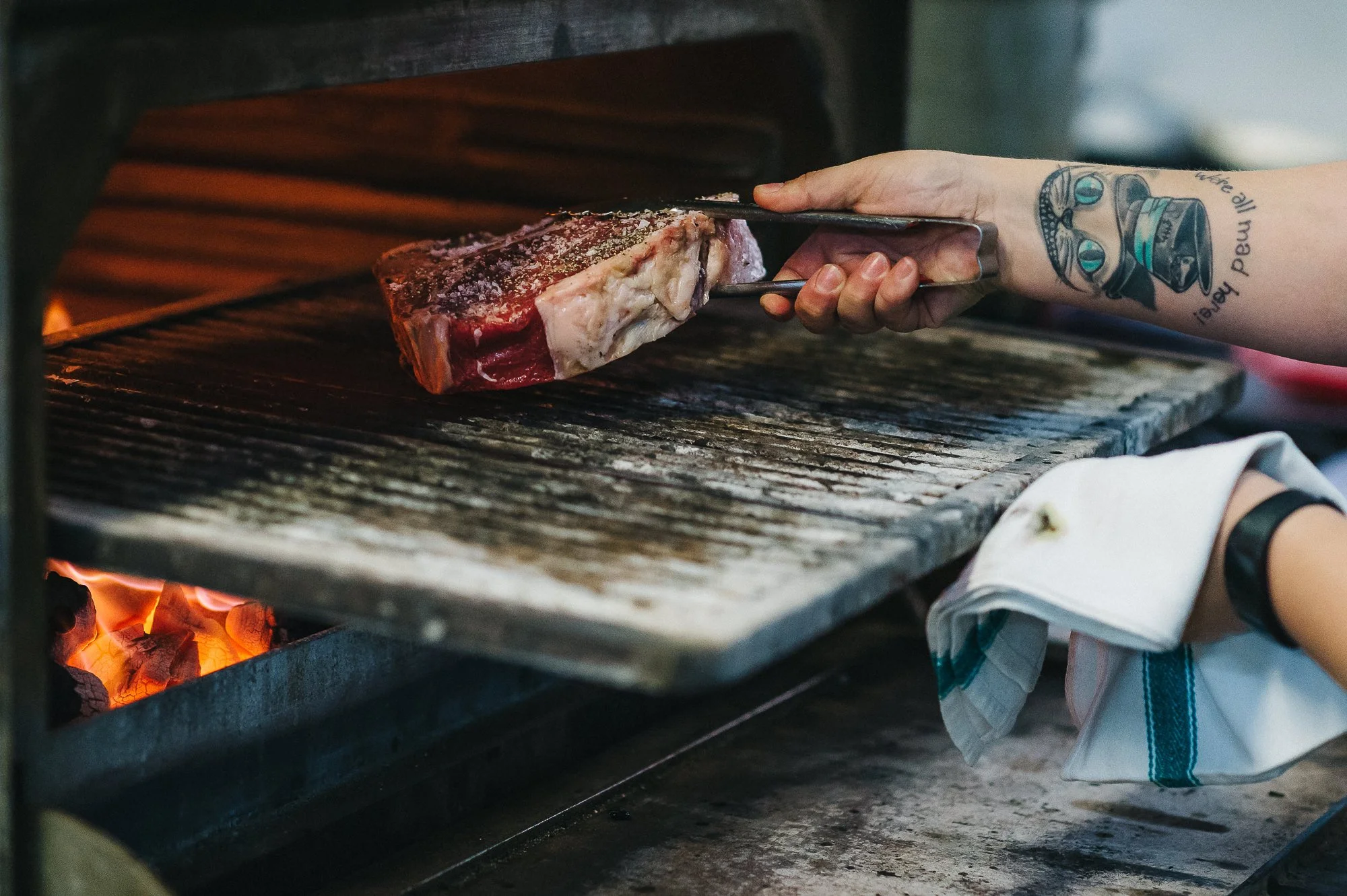 Person using tongs to place a large cut of raw meat into an open wood-fired grill.