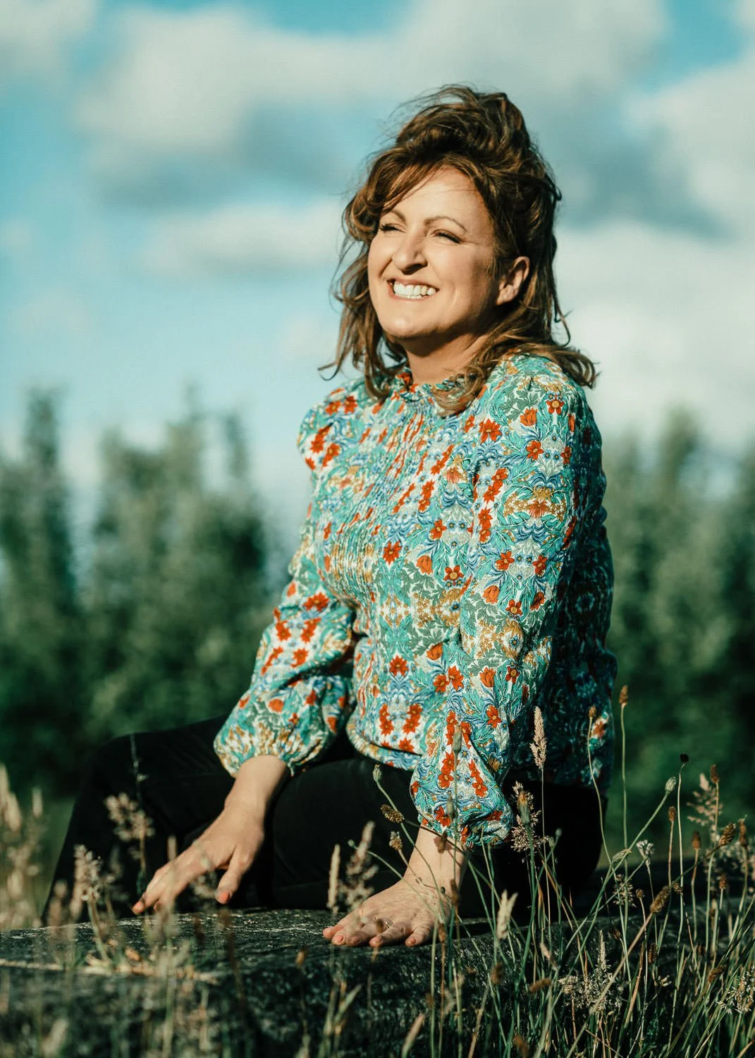 A woman with brown hair smiling and squatting outdoors in a field with tall grass, wearing a colorful floral blouse