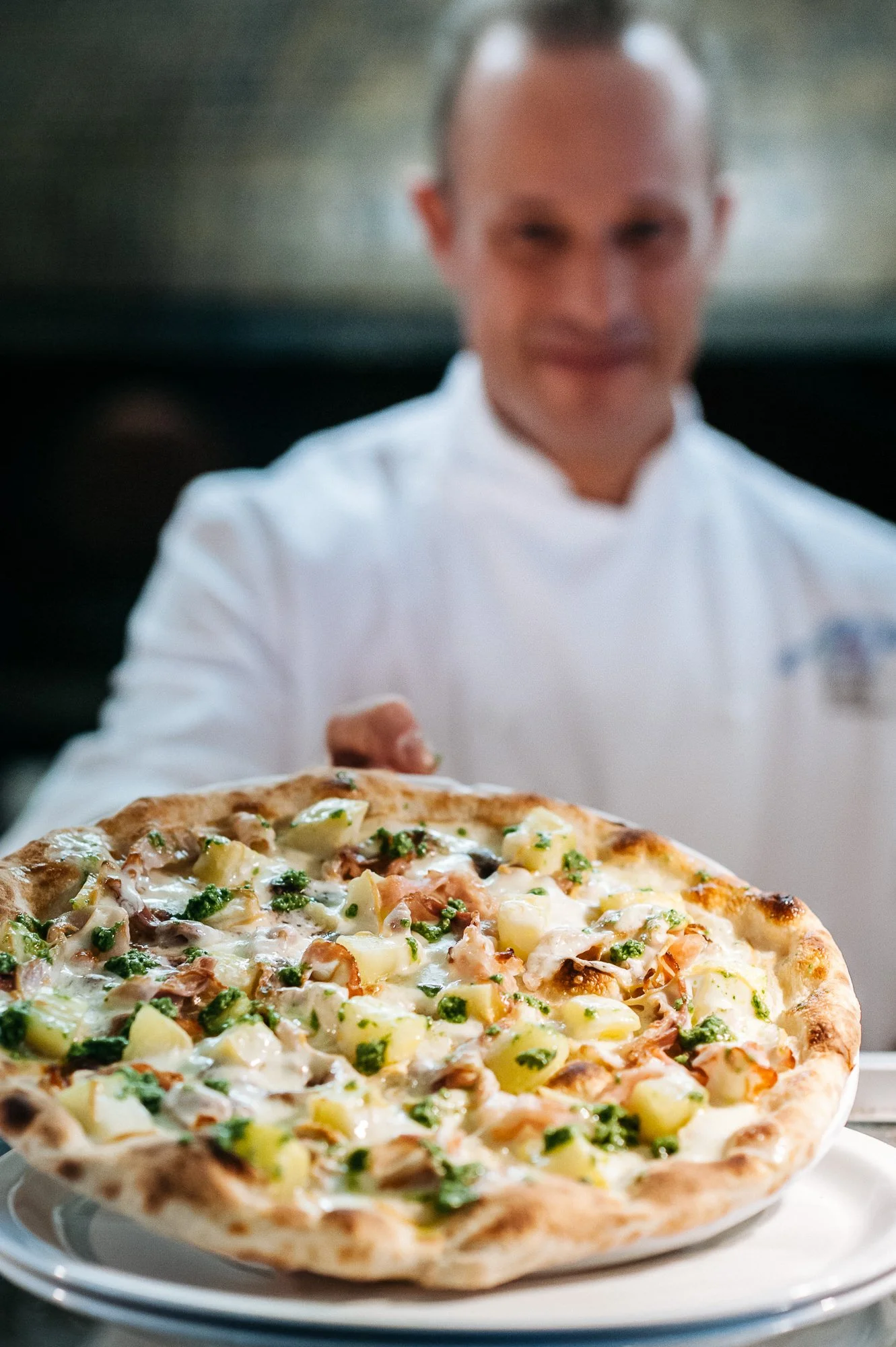 Chef placing a pizza topped with cheese, potatoes, green herbs, and possibly chicken in a restaurant kitchen.