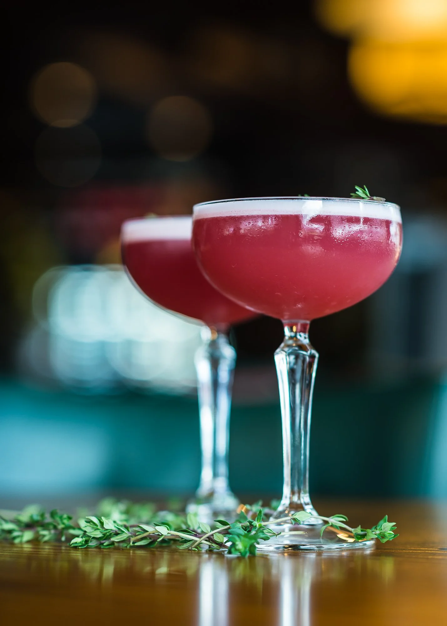 Two pink cocktails in elegant stemmed glasses garnished with small green herbs, placed on a wooden surface with a few green herbs at the base, with a blurred background of colorful lights.