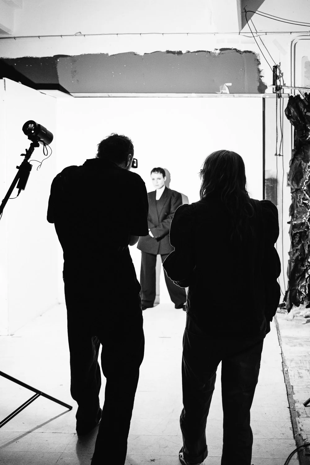 A black-and-white photo of a photoshoot studio. A model in a blazer poses in front of a white backdrop. Two people, likely crew members, are working nearby; one is taking photos while the other observes. Studio lighting equipment is set up, and the e