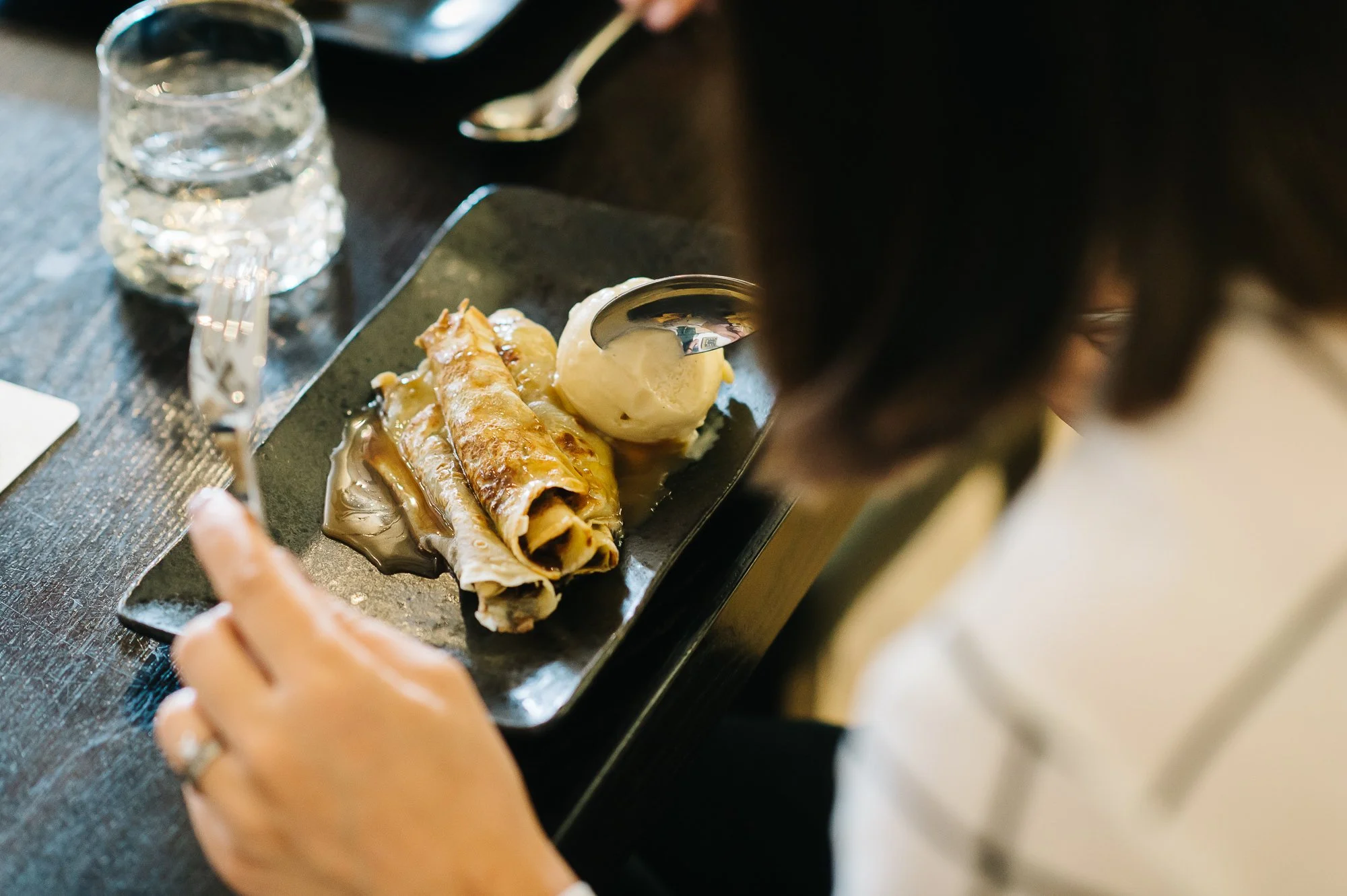 A person holding a fork and looking at a dish of cinnamon roll crepes with a scoop of vanilla ice cream on a black serving plate.