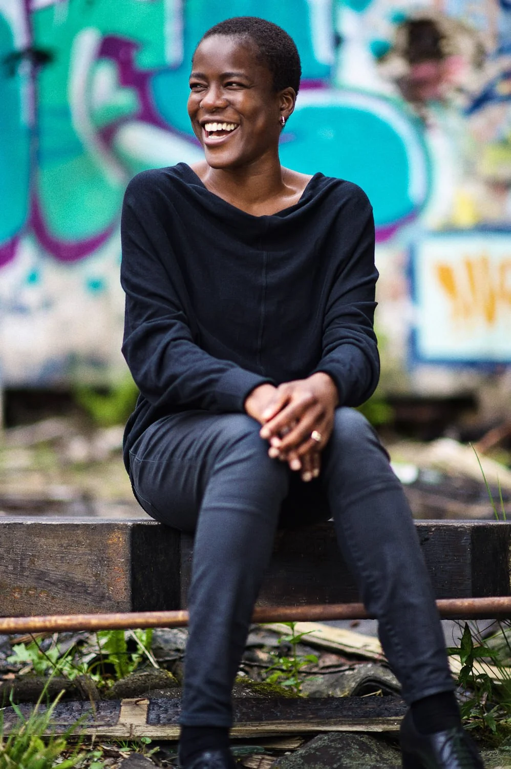 A woman with short hair, wearing a black hoodie and black pants, sitting on a wooden platform outdoors, laughing with a graffiti-covered wall in the background.