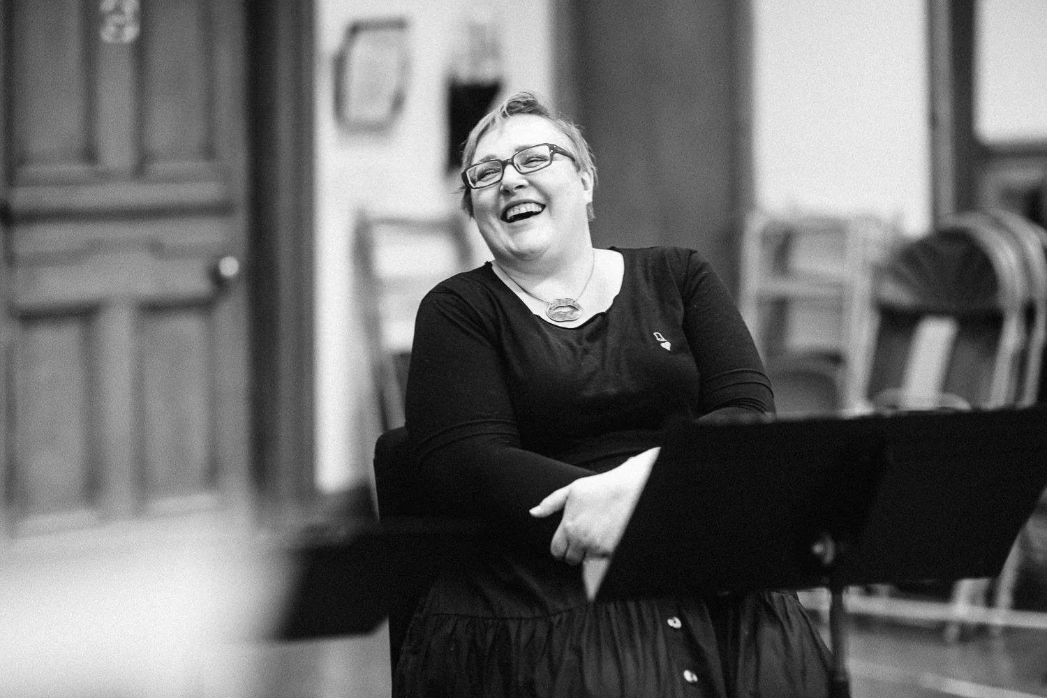A woman with glasses and short hair laughing, sitting in a room with wooden furniture.