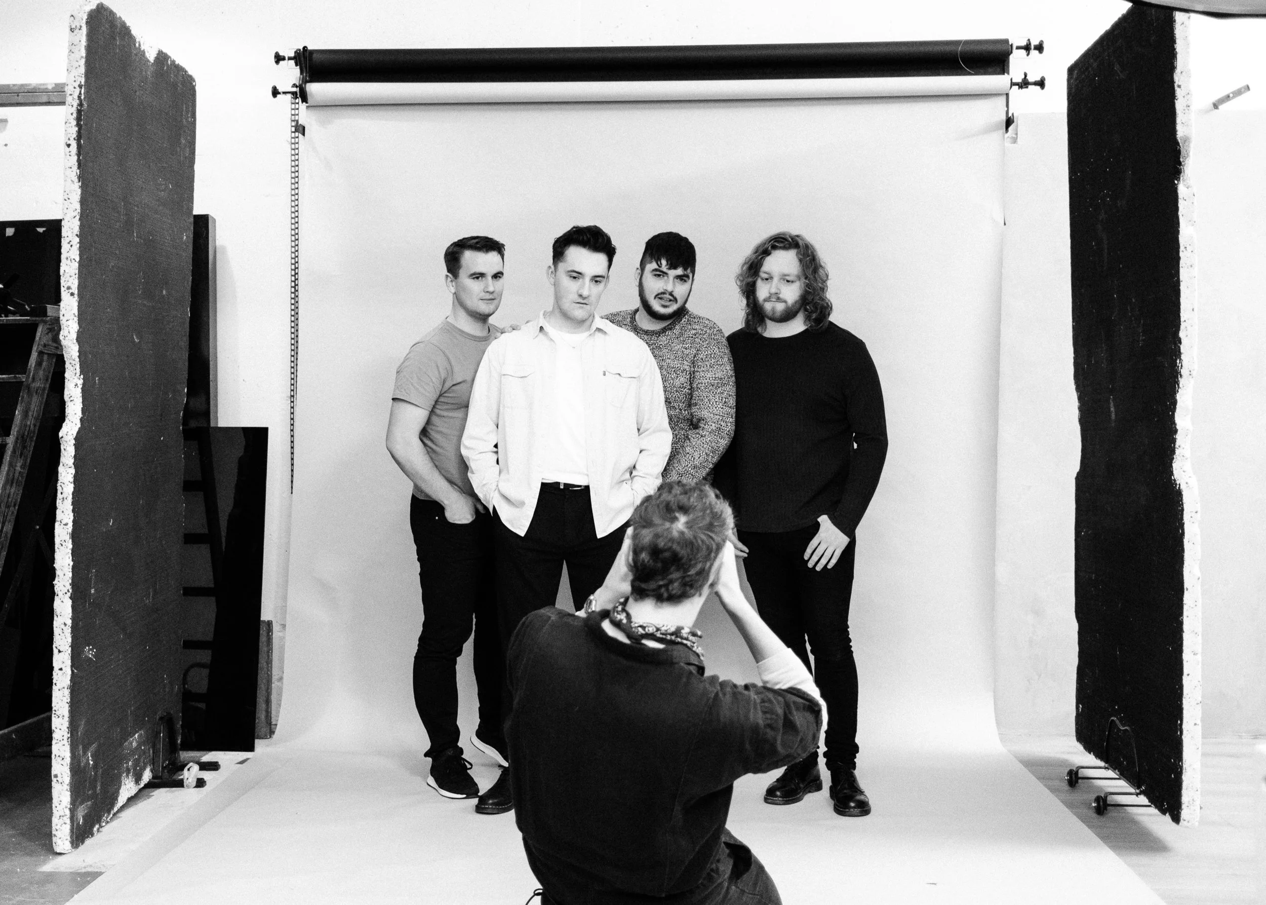 Black and white photo of four young men posing for a photo at a photography studio, with a photographer kneeling in front capturing the shot, in front of a plain backdrop with studio lighting and props visible.