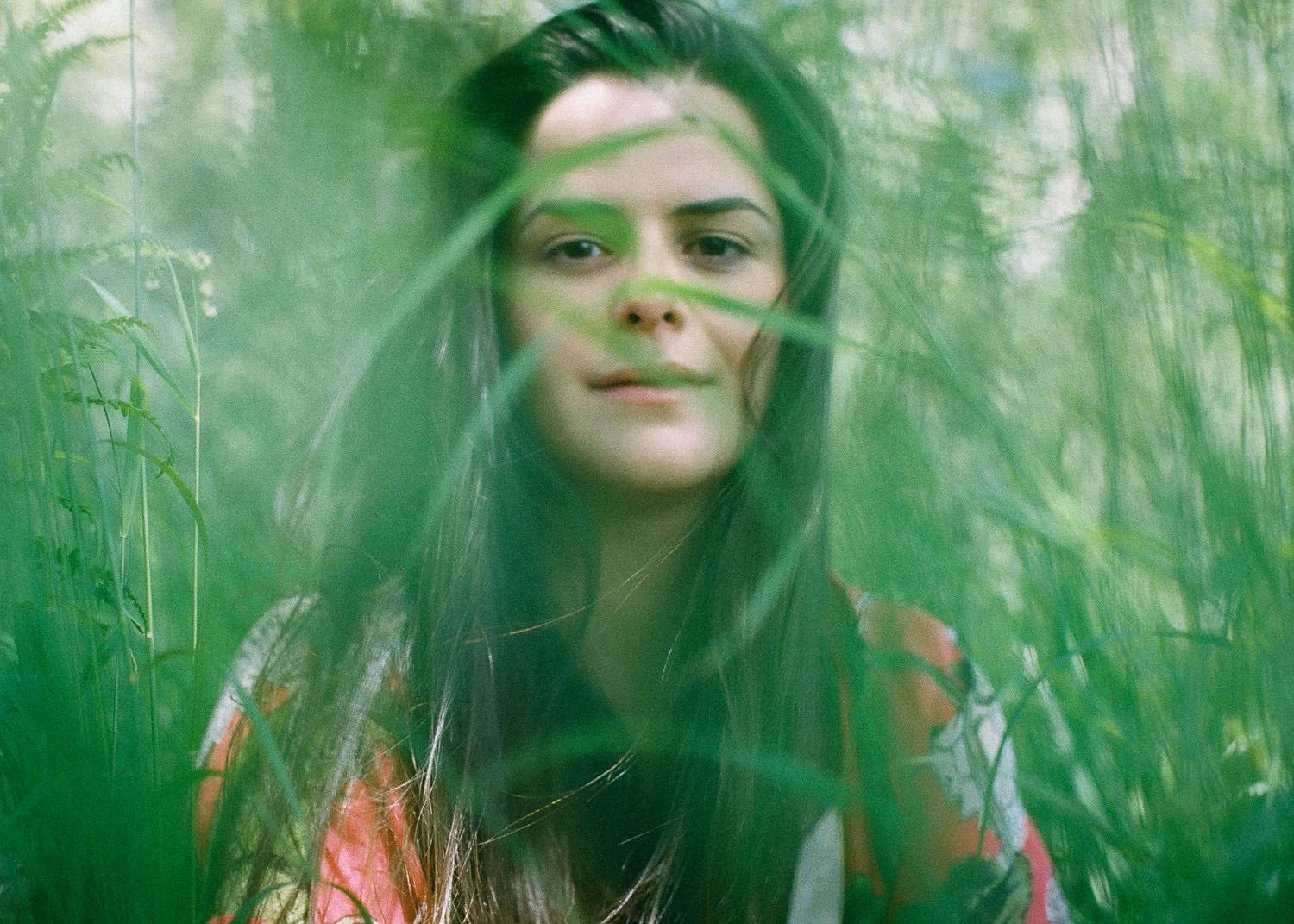 A woman with long dark hair and light skin looking through tall green grass in a natural outdoor setting.
