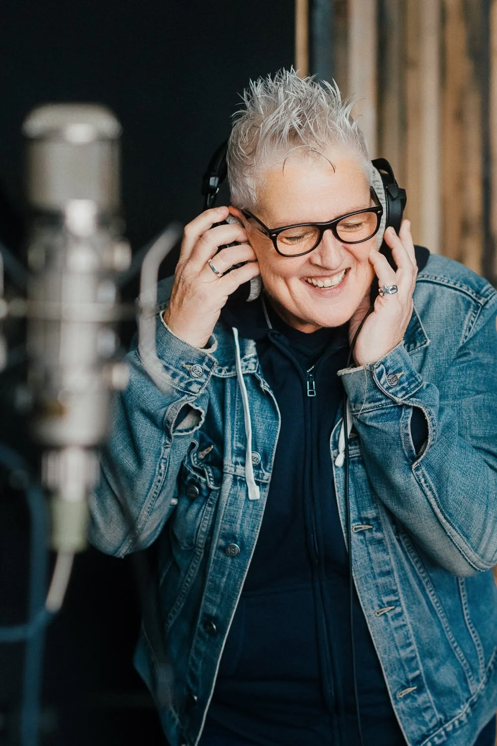 A person with short, spiky gray hair and glasses wearing a denim jacket and headphones, smiling while listening to music in a recording studio.