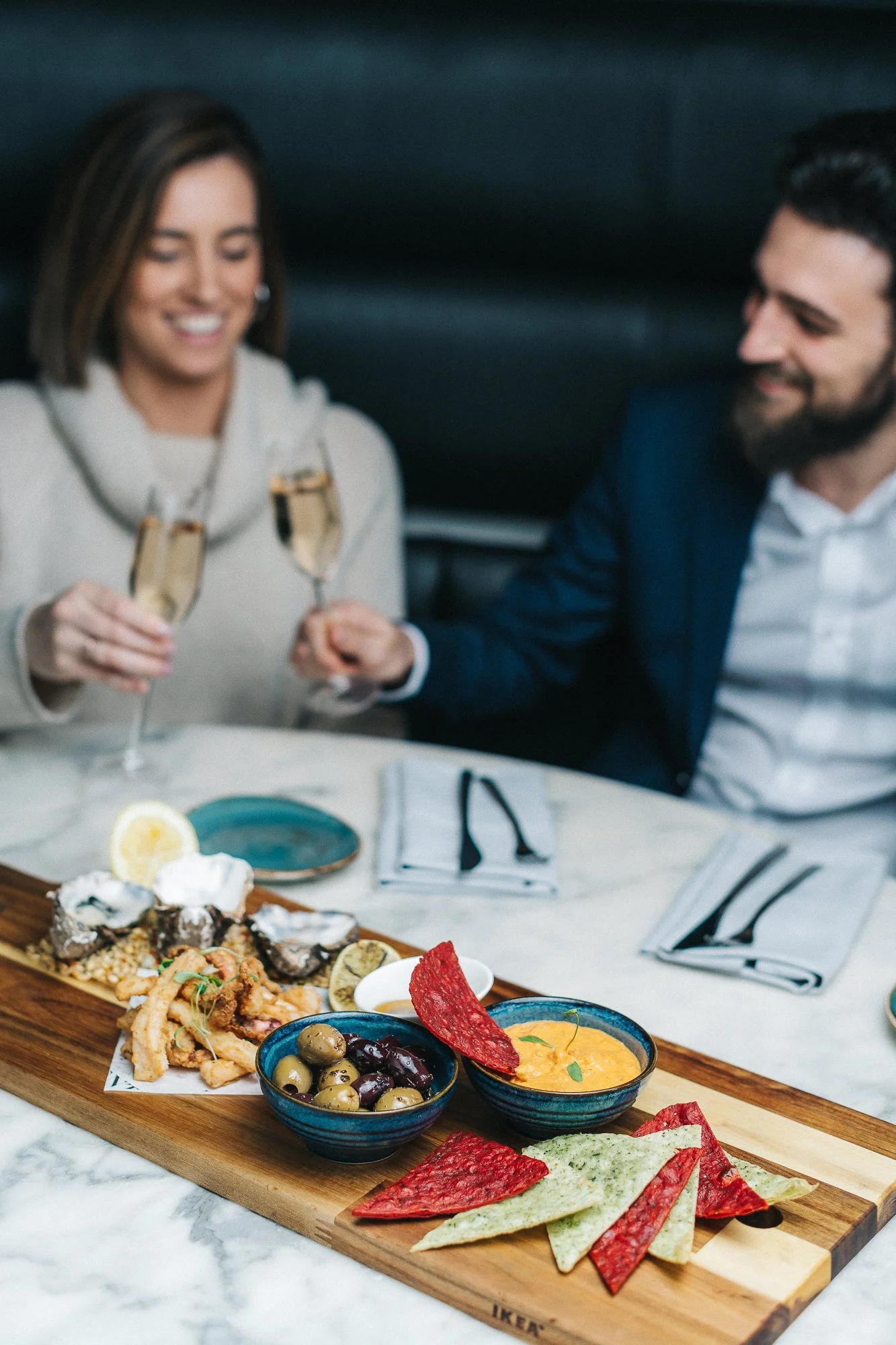 A man and a woman celebrating with champagne glasses at a table with a charcuterie board including olives, crackers, and colorful chips, in a restaurant setting.