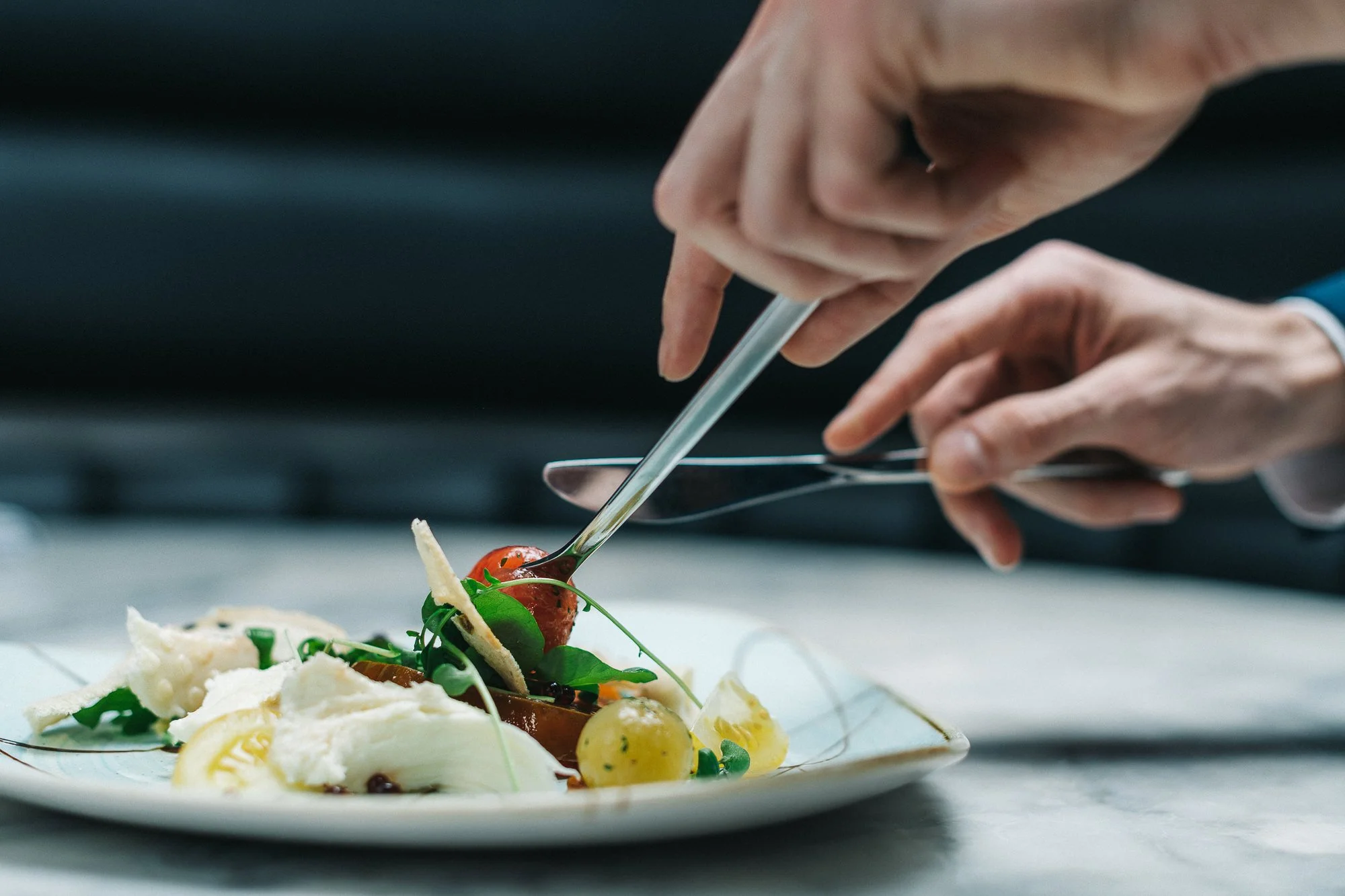 Close-up of a chef's hands arranging a gourmet dish on a white plate, using tweezers.