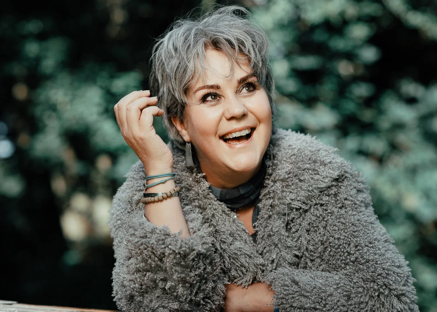 A joyful older woman with short gray hair and expressive eyes, wearing a cozy gray fur coat, surrounded by a blurred lush green outdoor background, smiling and looking up.
