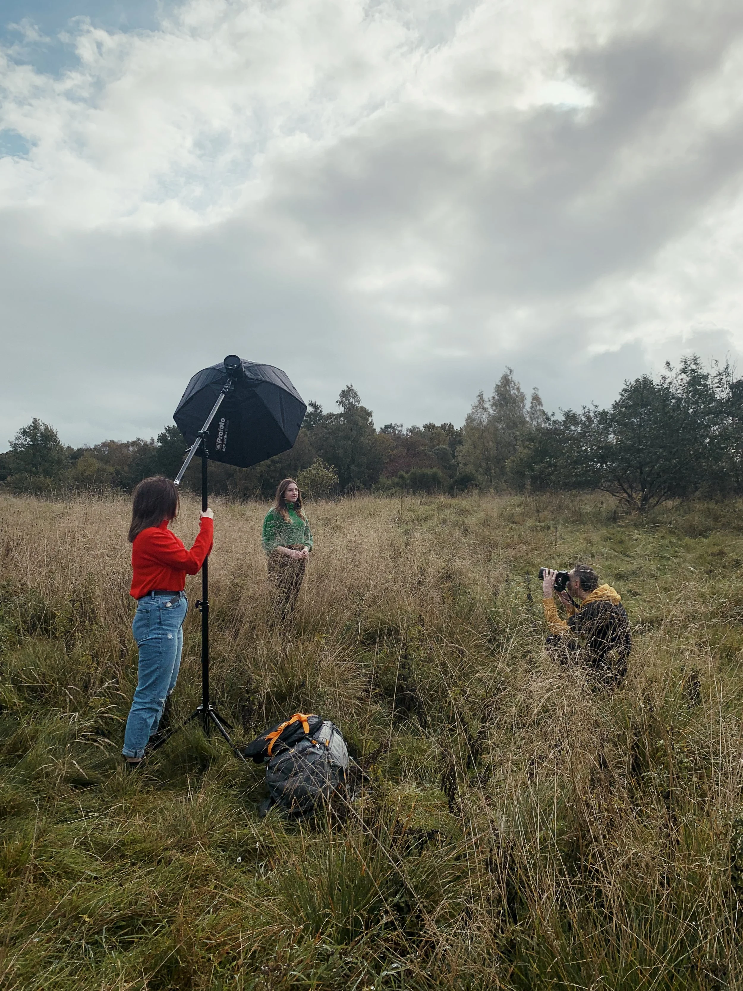 A woman in a green shirt is being photographed in a grassy field by a photographer kneeling in front of her. Another woman in a red jacket is holding an umbrella and standing nearby. A camera bag is on the ground in front of the woman with the umbrel