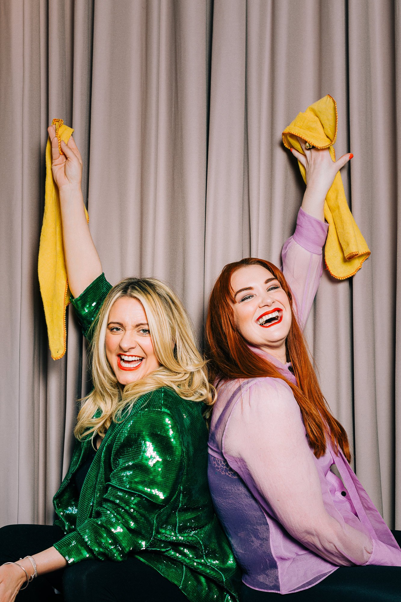 Two women sitting back to back, smiling and laughing, holding yellow cloths up in the air, with gray curtains in the background.
