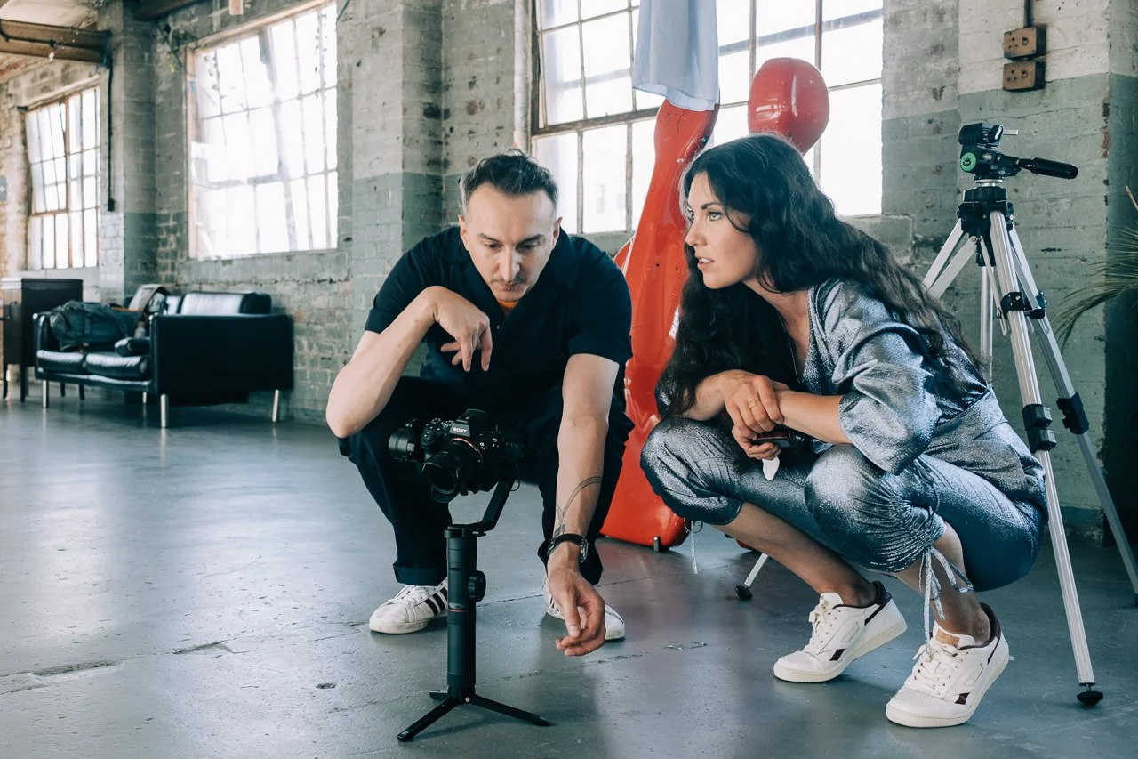 Two people, a man and a woman, crouch on a concrete floor in a spacious room with brick walls, examining a camera mounted on a tripod. The room has large windows, a black leather couch, a red punching bag, and a telescope.