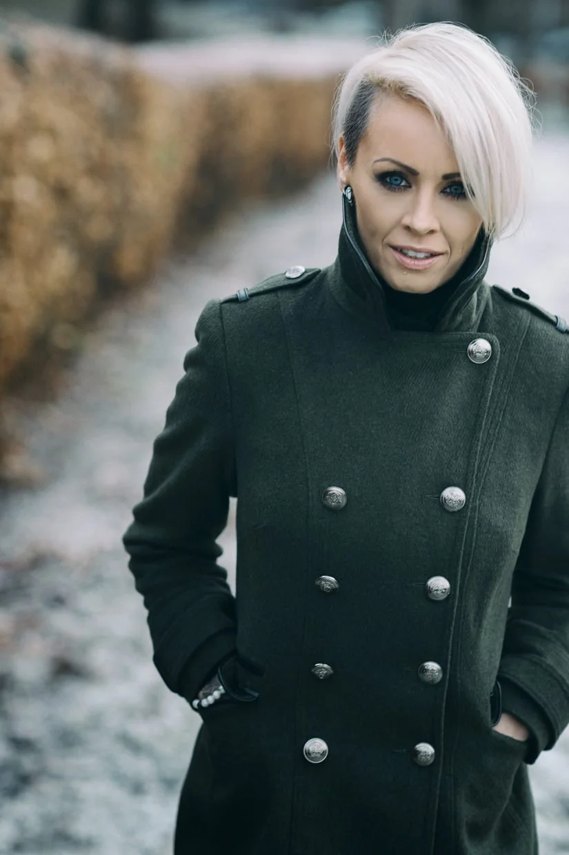 Woman with platinum blonde hair and blue eyes wearing a dark green military-style coat with silver buttons, outdoors near water and trees.