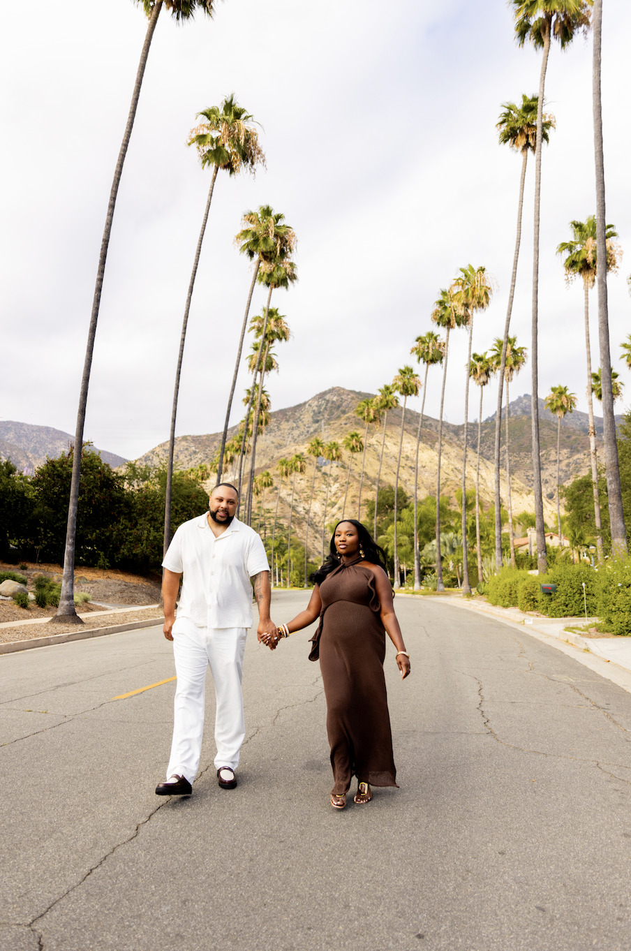 An expectant couple holds hands and walks down a palm-tree lined street in Southern California. There are mountains in the background. 