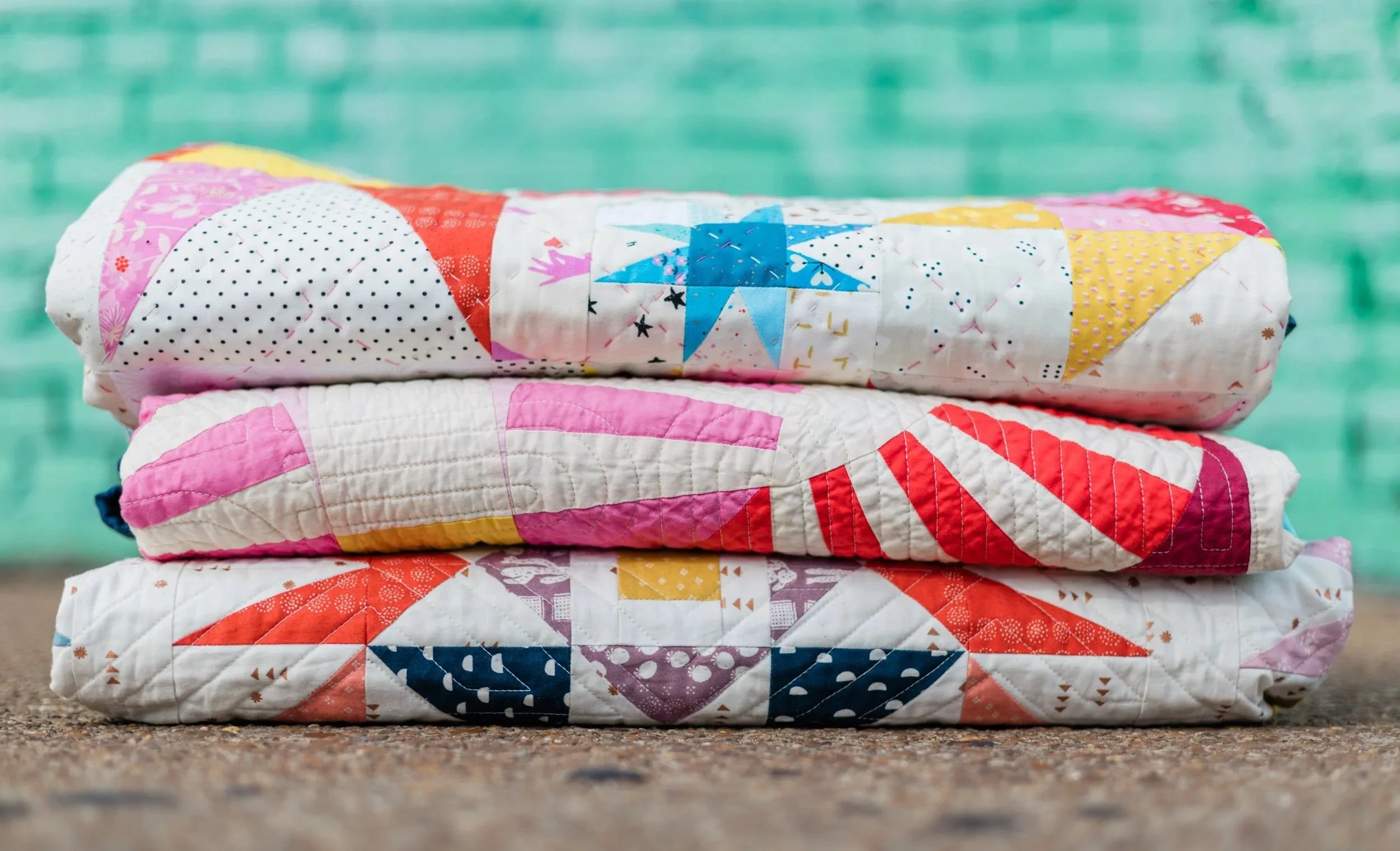 stack of She Makes Joy quilts folded and stacked in front of a green brick wall