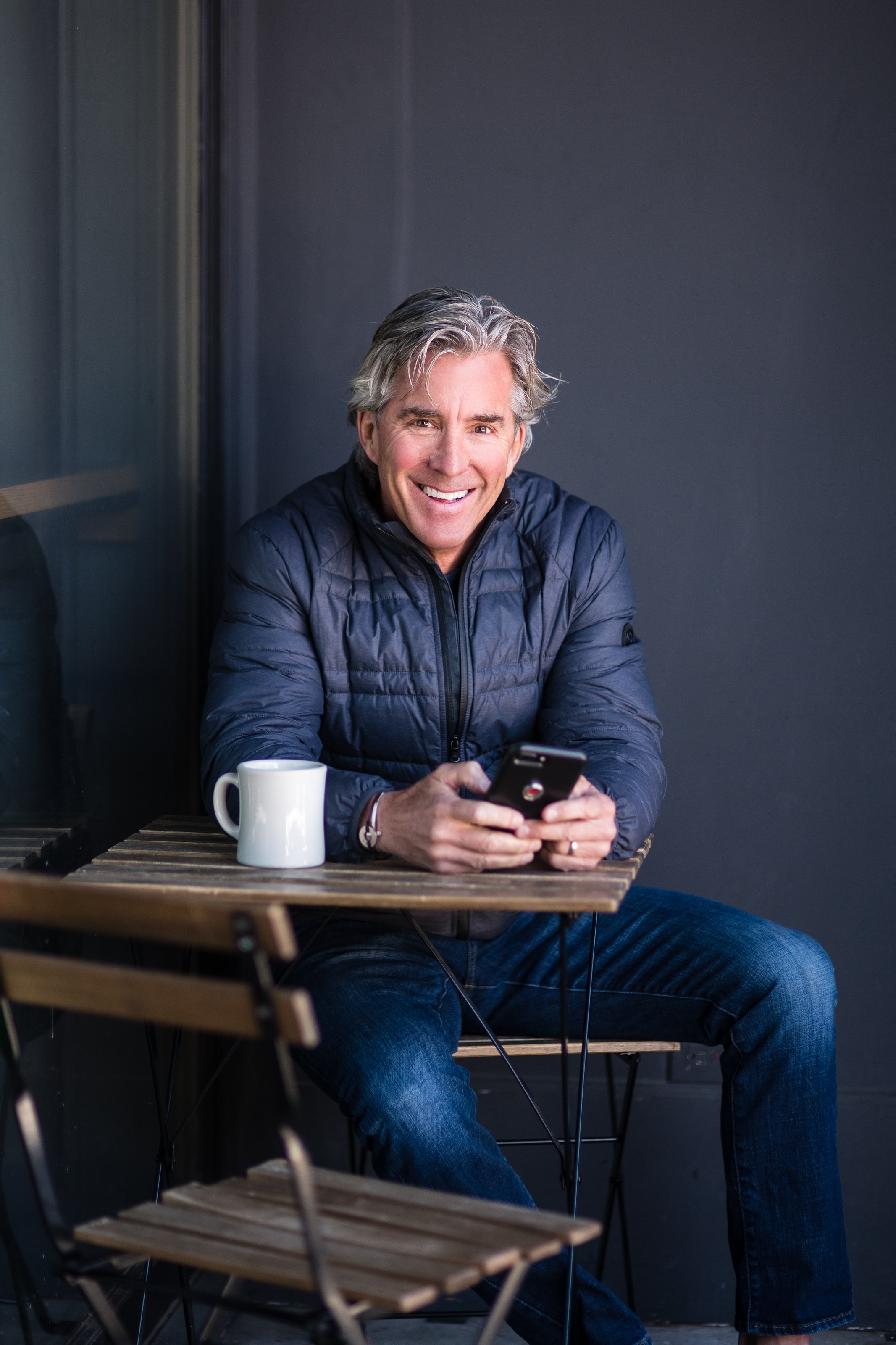 An older man with gray hair is sitting at a small wooden table, smiling and looking at his smartphone. He has a white mug on the table and is wearing a dark puffy jacket and jeans.