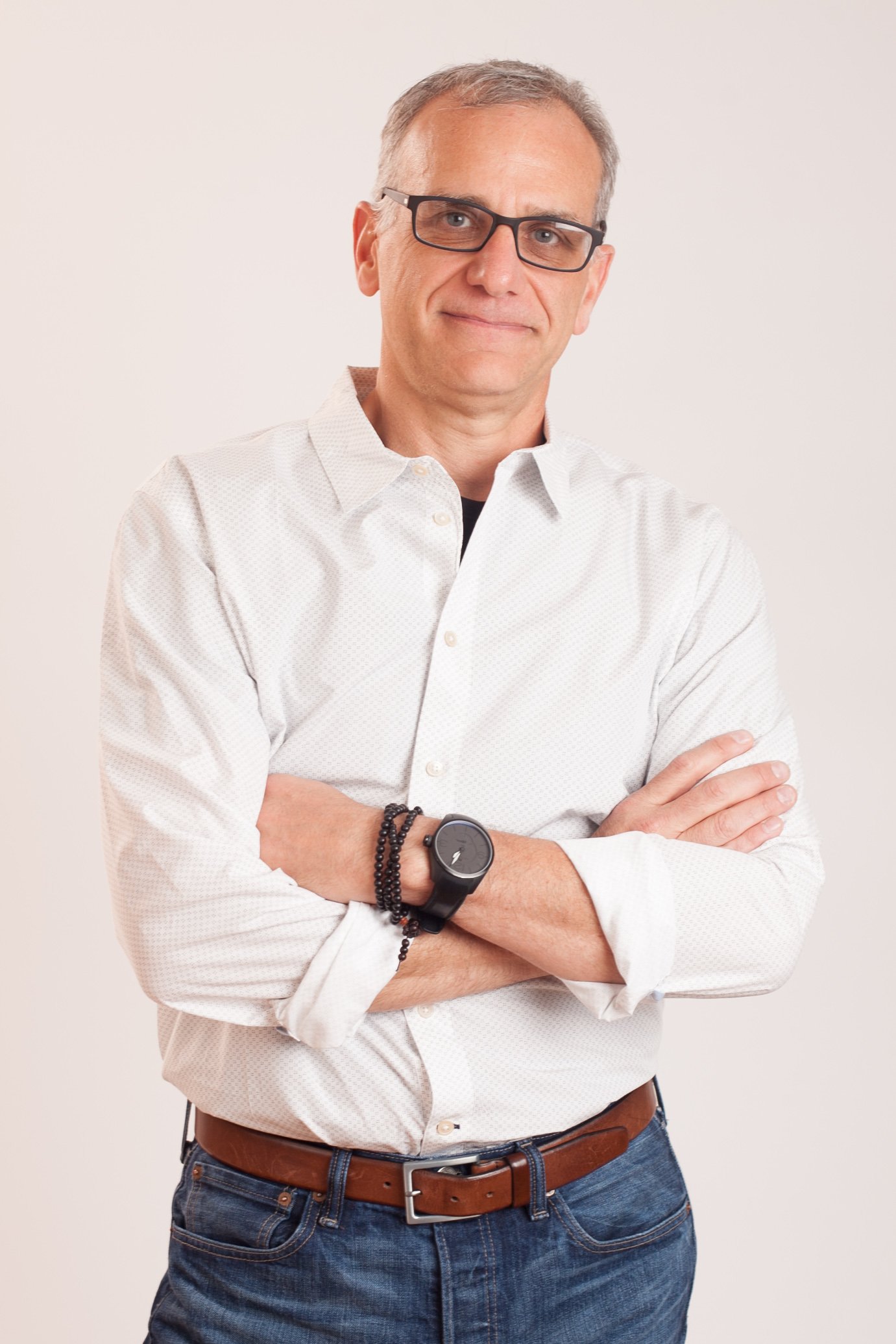 Portrait of a middle-aged man wearing glasses, a white button-up shirt with rolled-up sleeves, blue jeans, and a brown belt, standing with arms crossed against a plain light background.