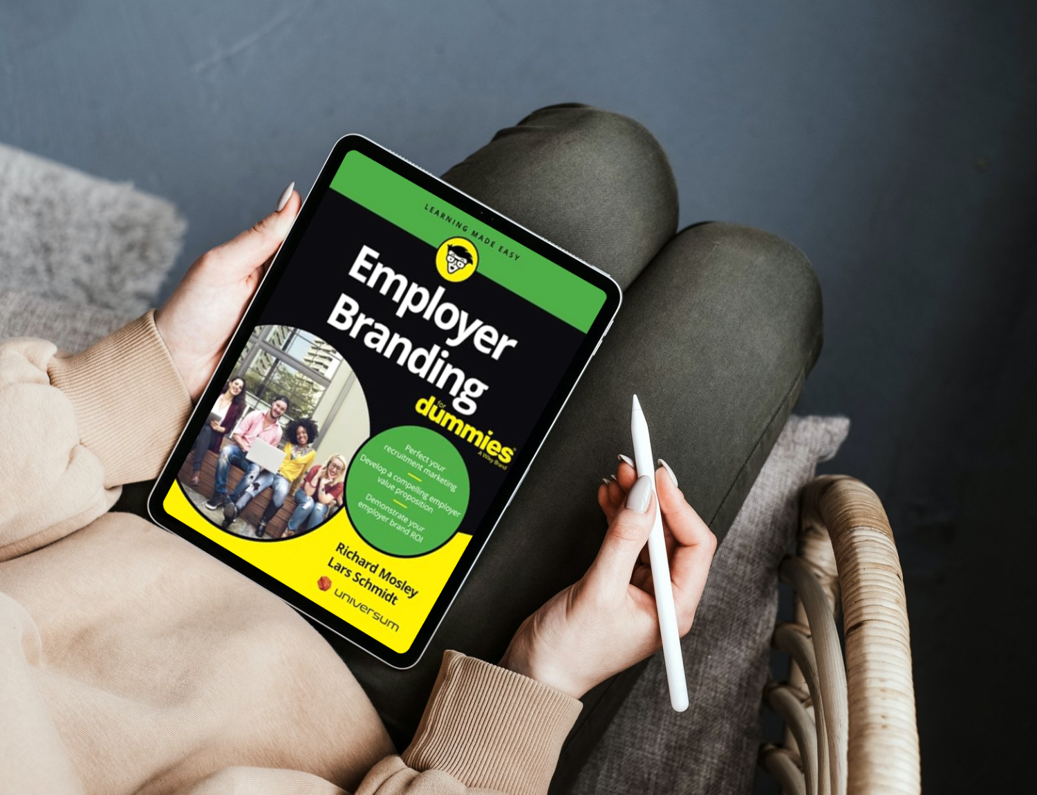 Person sitting on a chair holding a tablet displaying a book titled "Employer Branding for Dummies" with a photo of diverse people and the authors' names.