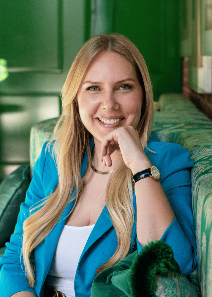 A woman with long blonde hair, smiling and wearing a blue blazer, sitting on a green patterned sofa in a room with brick and green walls.