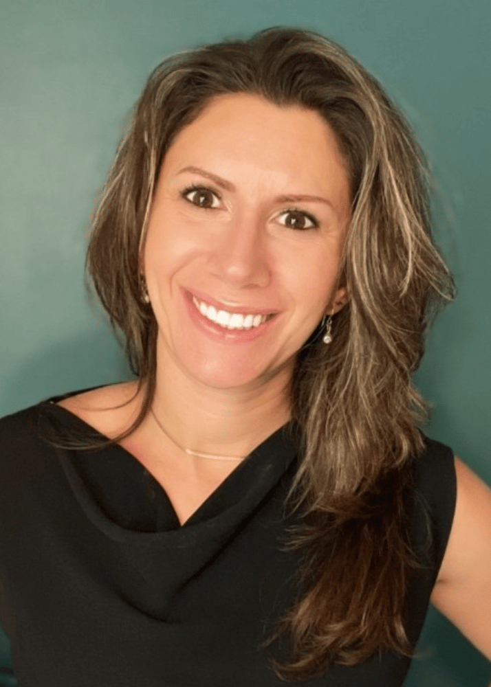 A woman with shoulder-length wavy brown hair smiling, wearing a black sleeveless top and a delicate necklace, standing against a teal background.