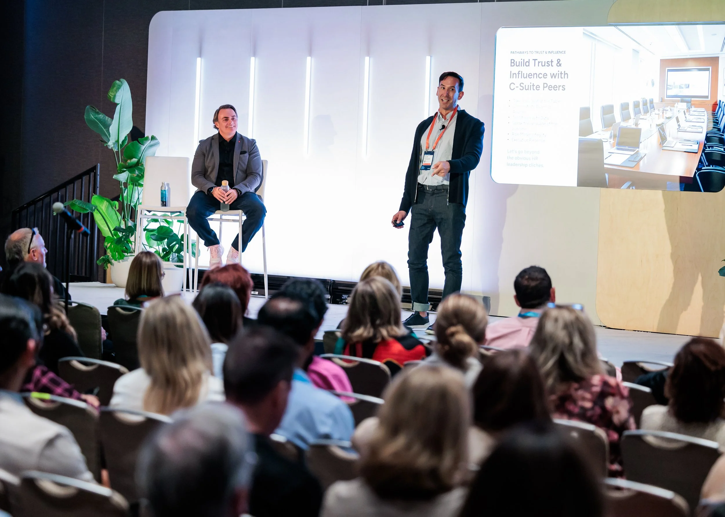 A man stands on stage giving a presentation to an audience seated in front of him. Another person sits on a chair on stage. A large screen displays a slide about building trust and influence with C-Suite peers.