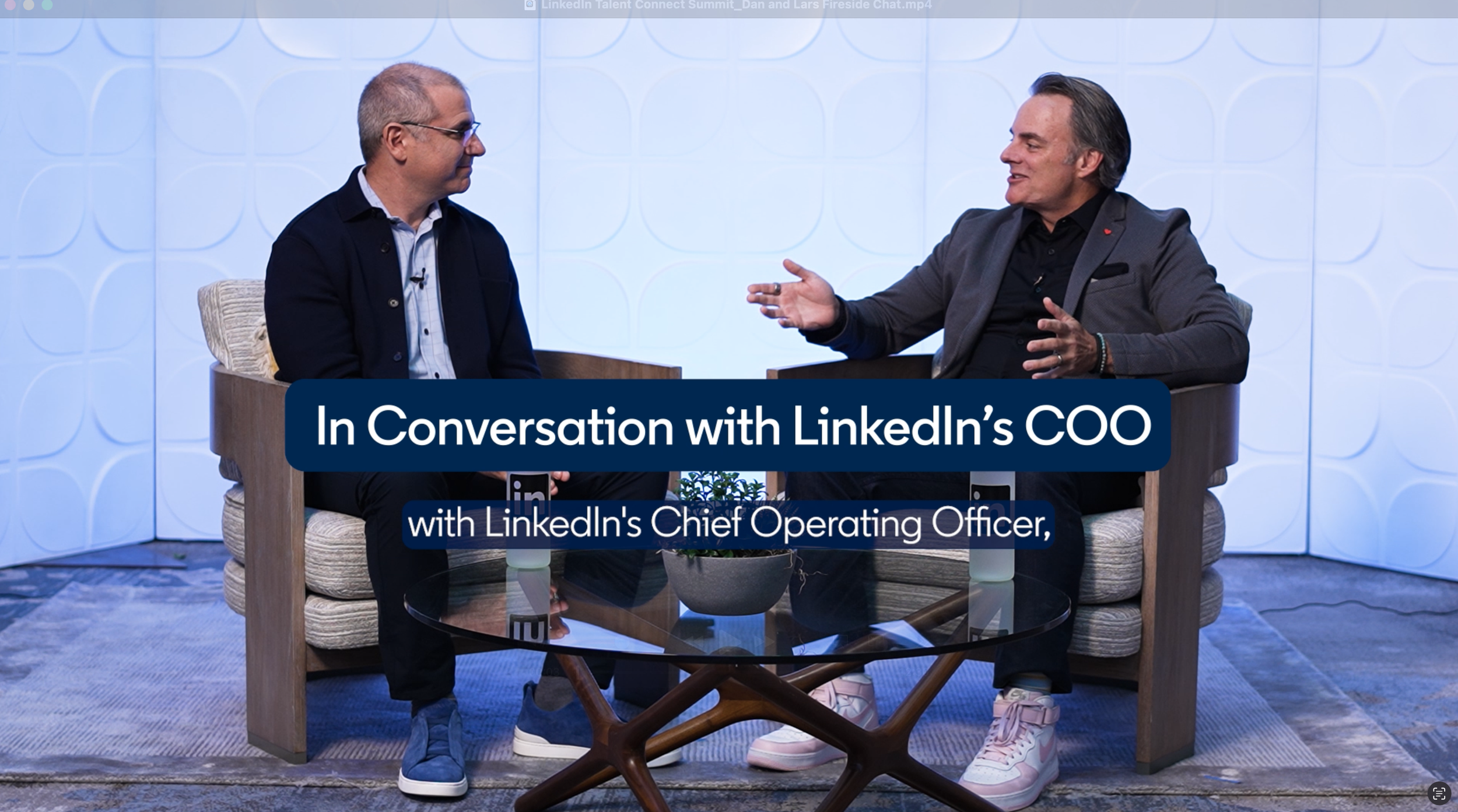 Two men sitting on armchairs in a studio, engaged in a conversation. One is wearing a dark blazer and jeans, and the other is wearing a suit jacket and sneakers. There is a glass-top table with a small plant in between them. The background is a light