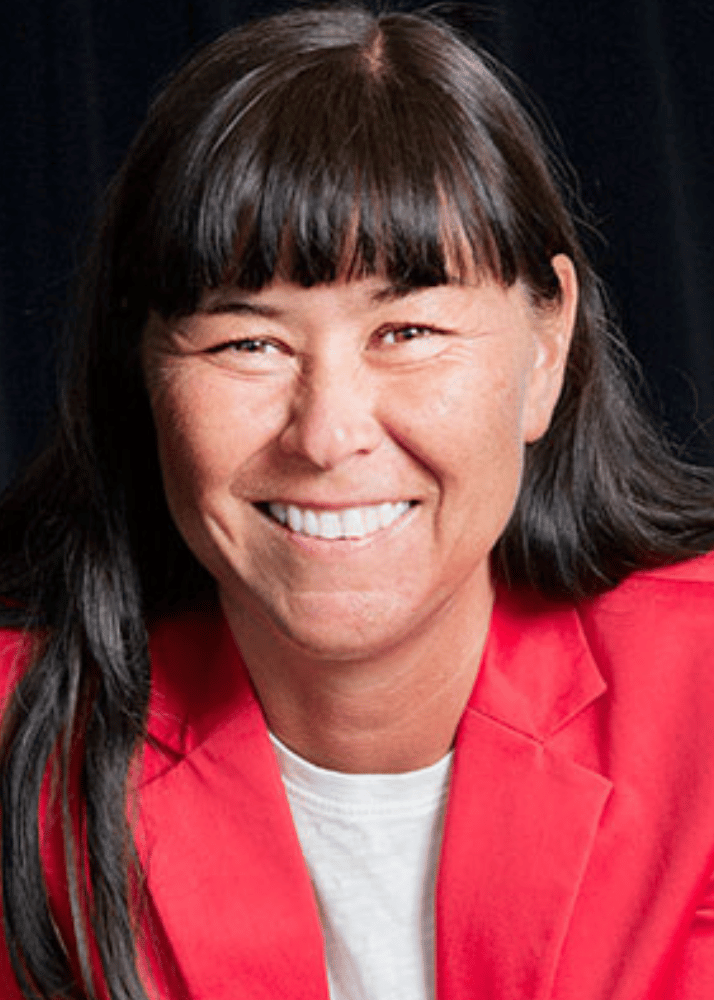 A woman with dark hair and bangs smiling in an indoor setting with plants in the background.