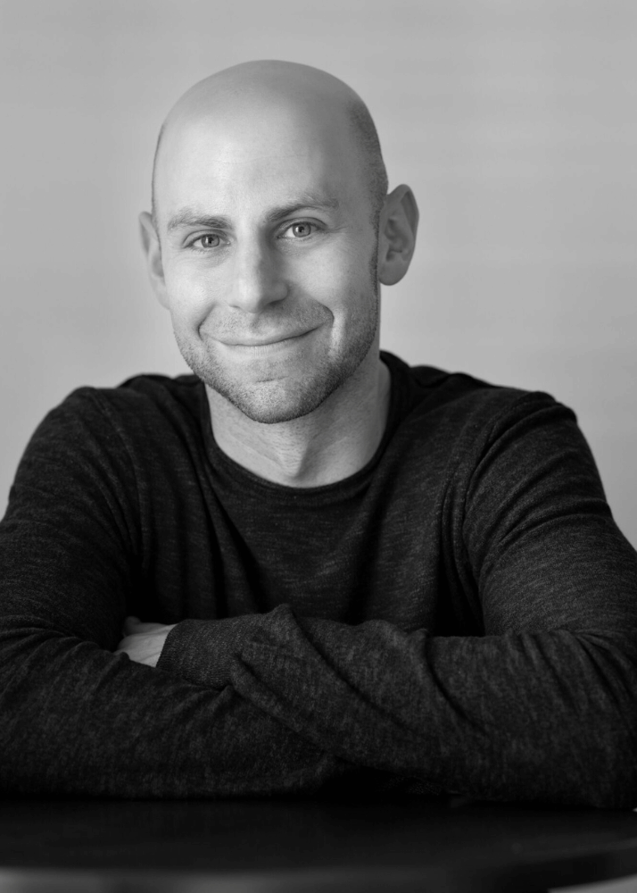 Black and white photo of a smiling bald man with light-colored eyes, wearing a dark long-sleeve shirt, sitting at a round table with his arms crossed, against a plain background.