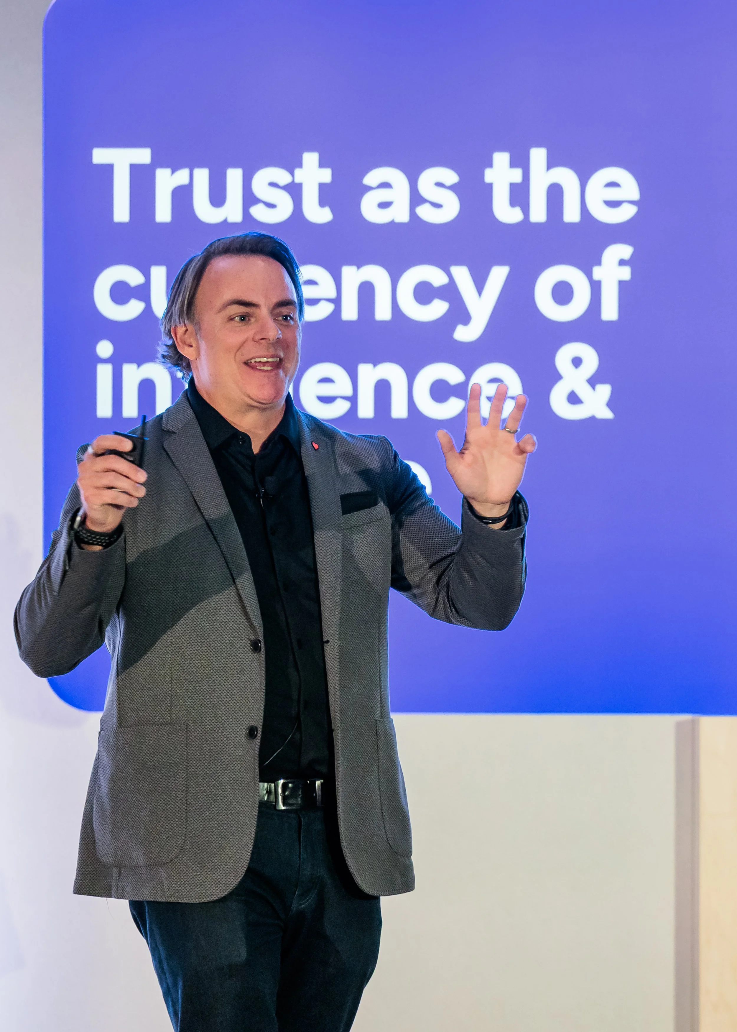 A man in a gray blazer and black shirt giving a presentation in front of a blue slide that reads, 'Trust as the currency of influence &'.