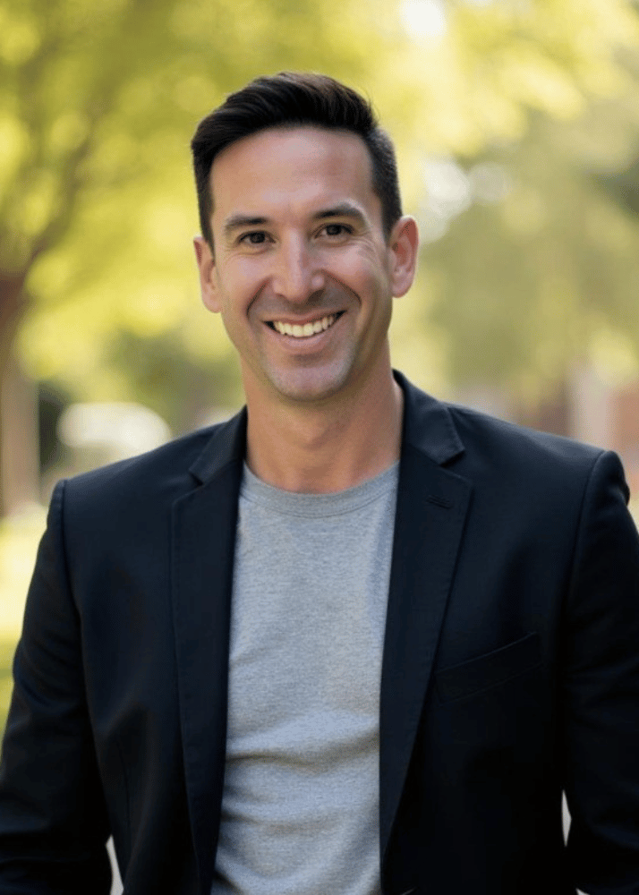A smiling man with dark hair wearing a black blazer and gray t-shirt outdoors with green trees and sunlight in the background.