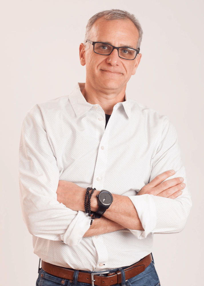 Portrait of a middle-aged man wearing glasses, a white button-up shirt with rolled-up sleeves, blue jeans, and a brown belt, standing with arms crossed against a plain light background.