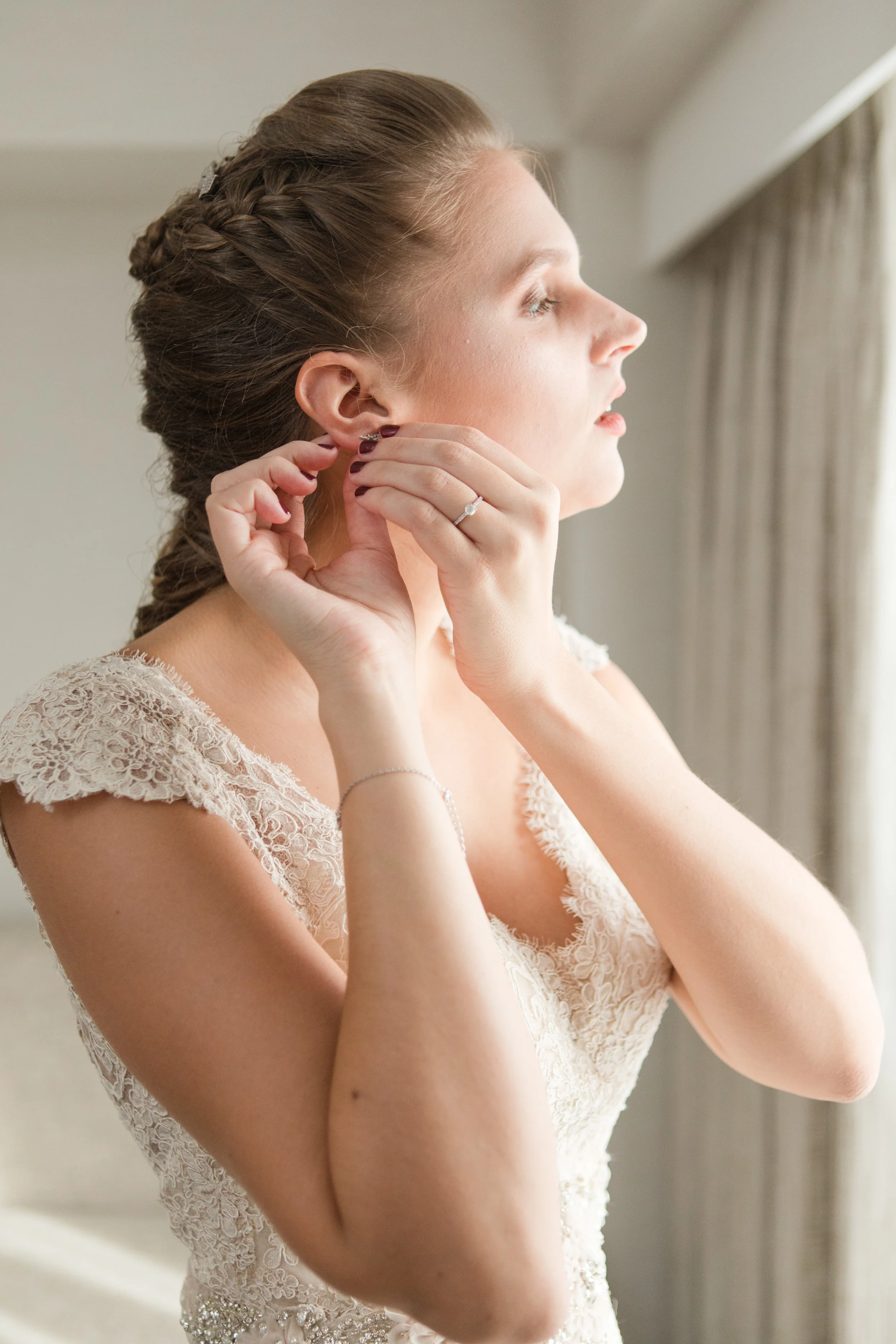bride putting in earrings on wedding day in lace allure bridal gown photographed by jaye kogut