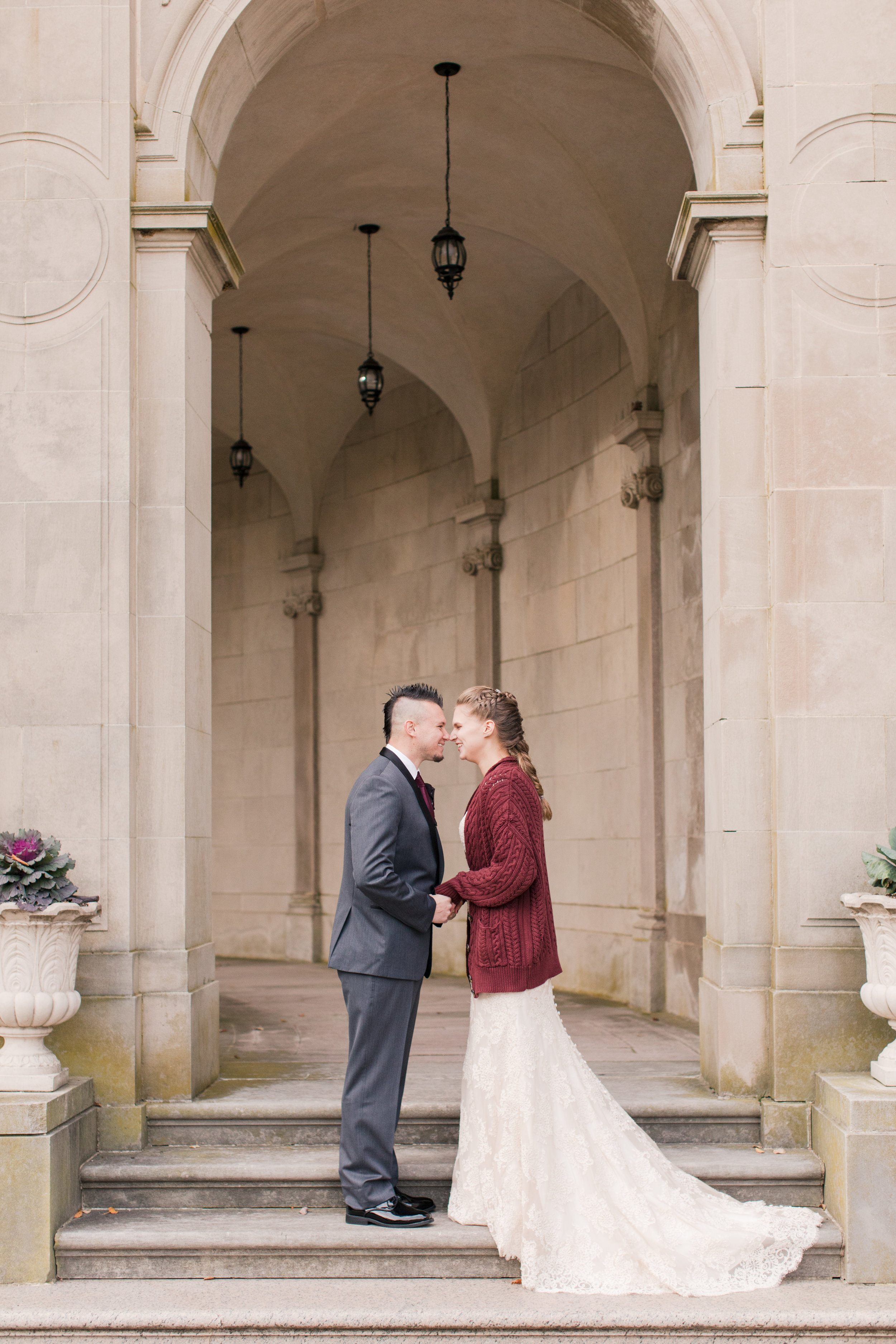 bride in lace allure bridal gown with buttons wearing chunky knit sweater stands with groom atop steps at monmouth university for bridal portraits