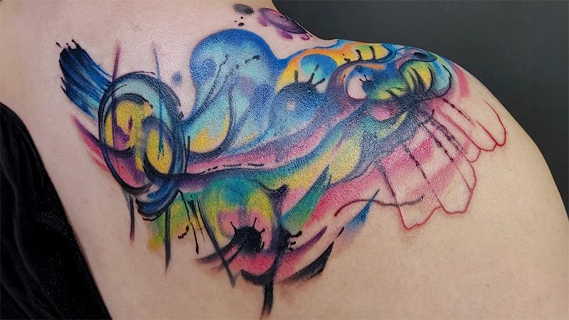 A colorful abstract tattoo on a womans sholder.  It features all colors of the rainbow with no distinct shapes or patterns.  It resembles colorful chaos, from a tattoo shop in Spokane, Washington.
