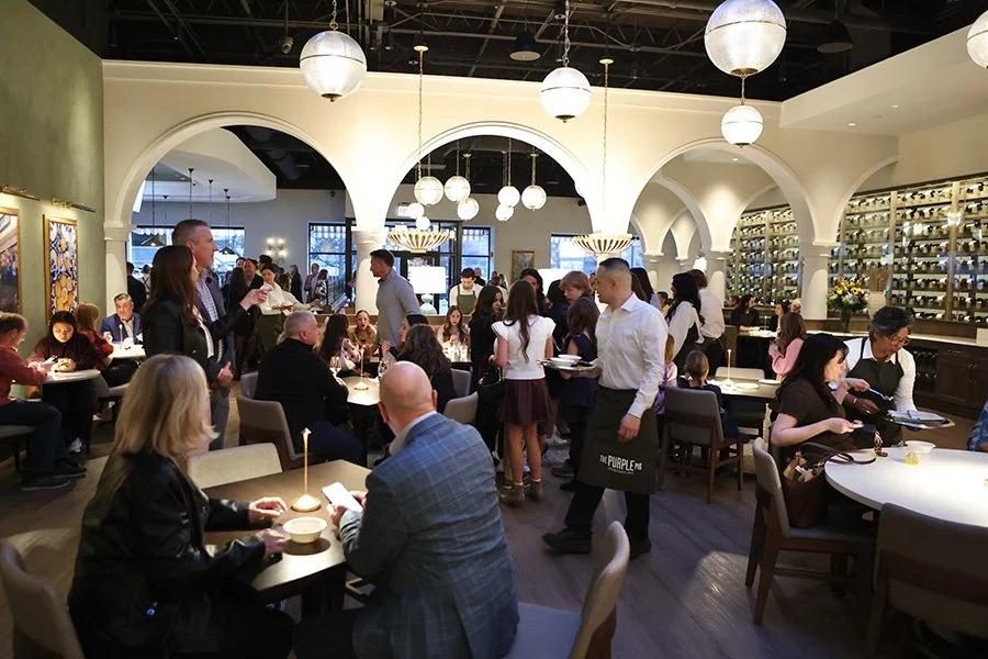 Busy restaurant with people celebrating the opening of The Purple Pig Oak Brook. Servers are serving food or small bites. The restaurant decor includes arches and white spherical pendant lights.