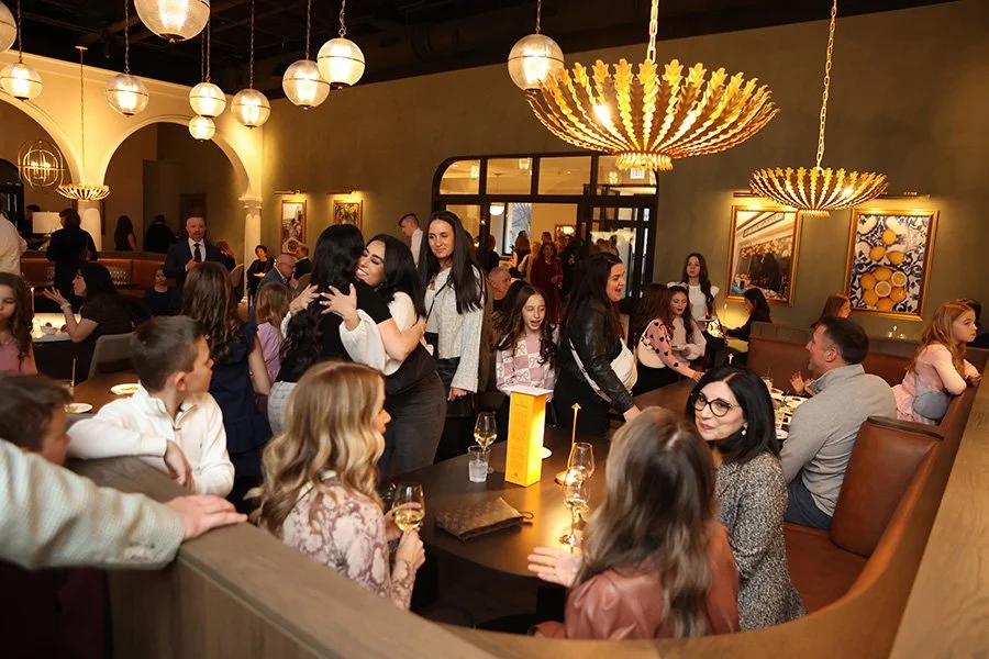 People celebrating the opening reception for The Purple Pig Oak Brook. Guests are sitting, standing, and hugging. There are gold chandeliers and artwork on the walls.