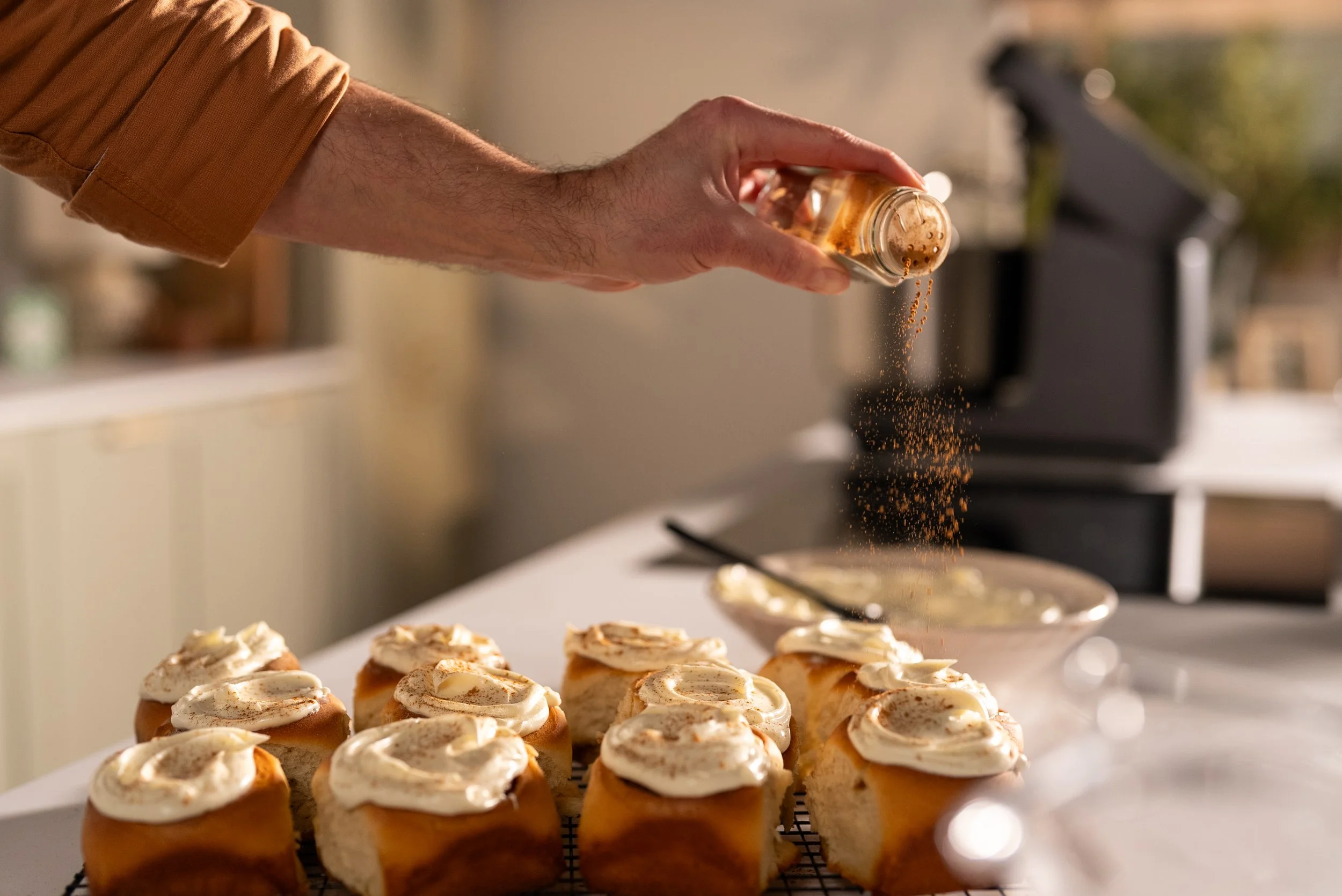 A person sprinkling cinnamon on a tray of frosted cupcakes in a kitchen.