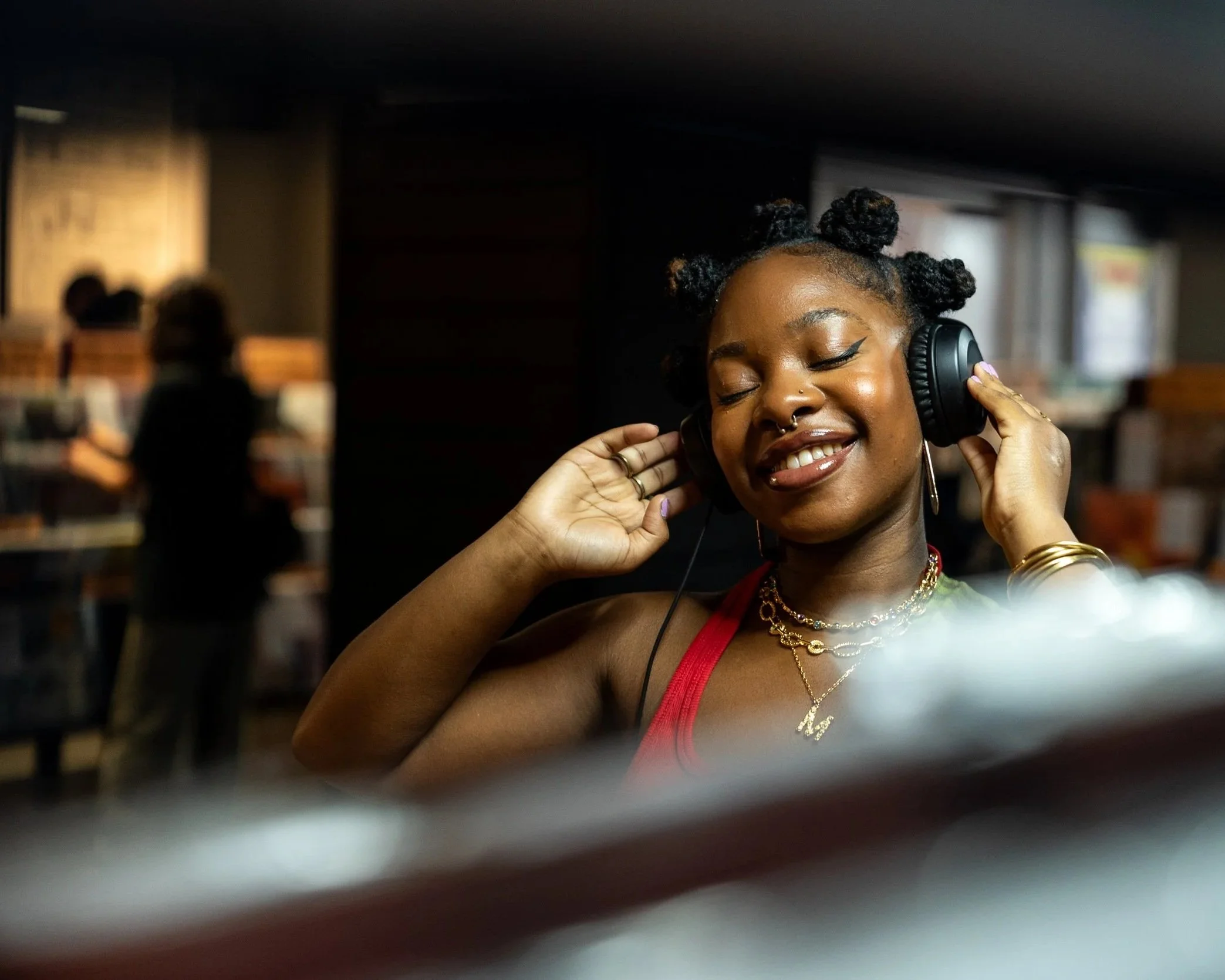 A woman with dark hair styled in bantu knots enjoying music with closed eyes, wearing headphones, jewelry, and a red top, in a cozy indoor setting.