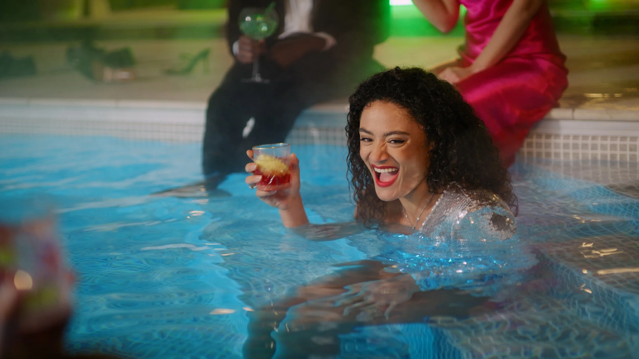 A woman with curly hair in a swimsuit is joyfully holding a drink while in a swimming pool, smiling and winking at the camera. Two other people are seen sitting by the poolside in the background.